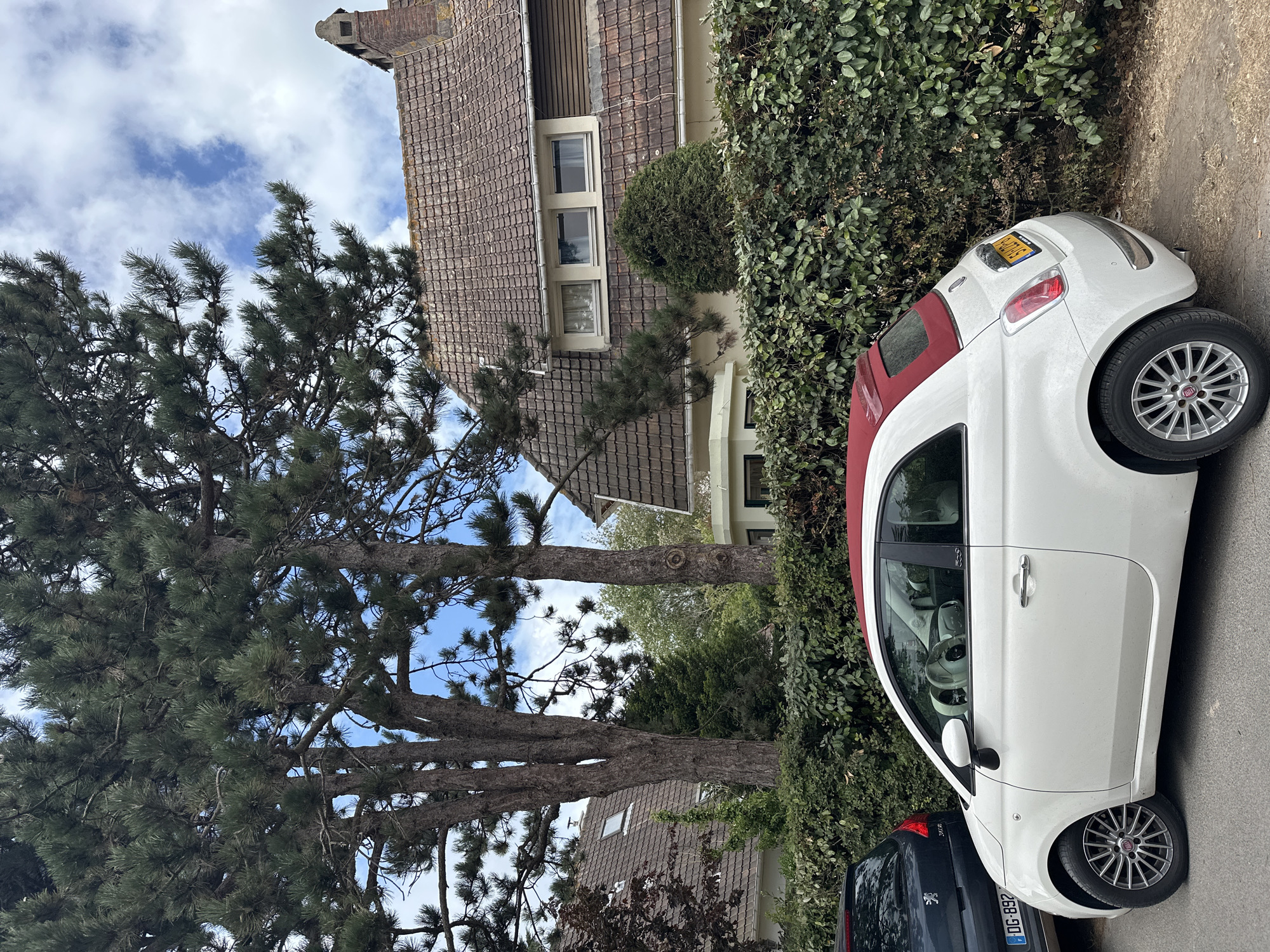 In Wissant, France, a white Fiat 500 convertible with a red soft top is parked on a street next to a dense green hedge. The car's left side is visible, showing its front wheel and the driver's side door. A dark blue Peugeot is parked to the left of the Fiat. Behind the hedge, a red-tiled roof house with several windows is visible, partially obscured by large trees with dark green needles. The sky above is blue with scattered white clouds.