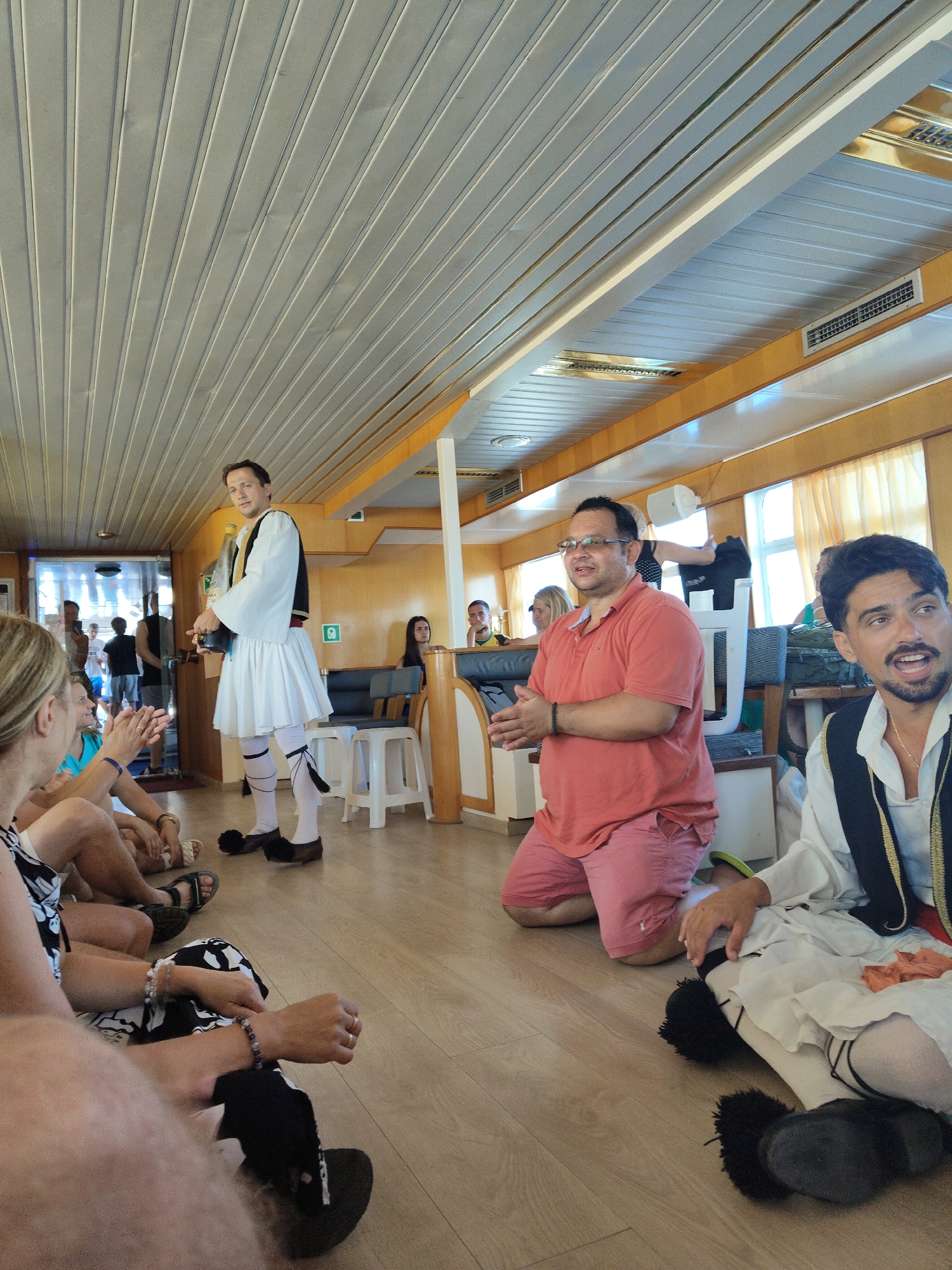 The image shows an indoor gathering on a boat, possibly a ferry, with people seated on the floor and on benches. A man dressed in traditional Greek attire, a white fustanella with black vest and gaiters, stands in the center of the image, holding a bottle and a small case. To his right, a man in a coral-colored polo shirt and pink shorts is kneeling. In the foreground, people are seated on the wooden floor, some with their backs to the camera. In the background, other passengers are visible, some looking towards the man in traditional dress. The environment appears to be a passenger area with cushioned benches, windows with yellow curtains, and a slatted ceiling. The overall scene suggests a cultural performance or demonstration for tourists. The location is not specified, but the traditional Greek clothing suggests it is somewhere in Greece, or a place with a significant Greek cultural presence.