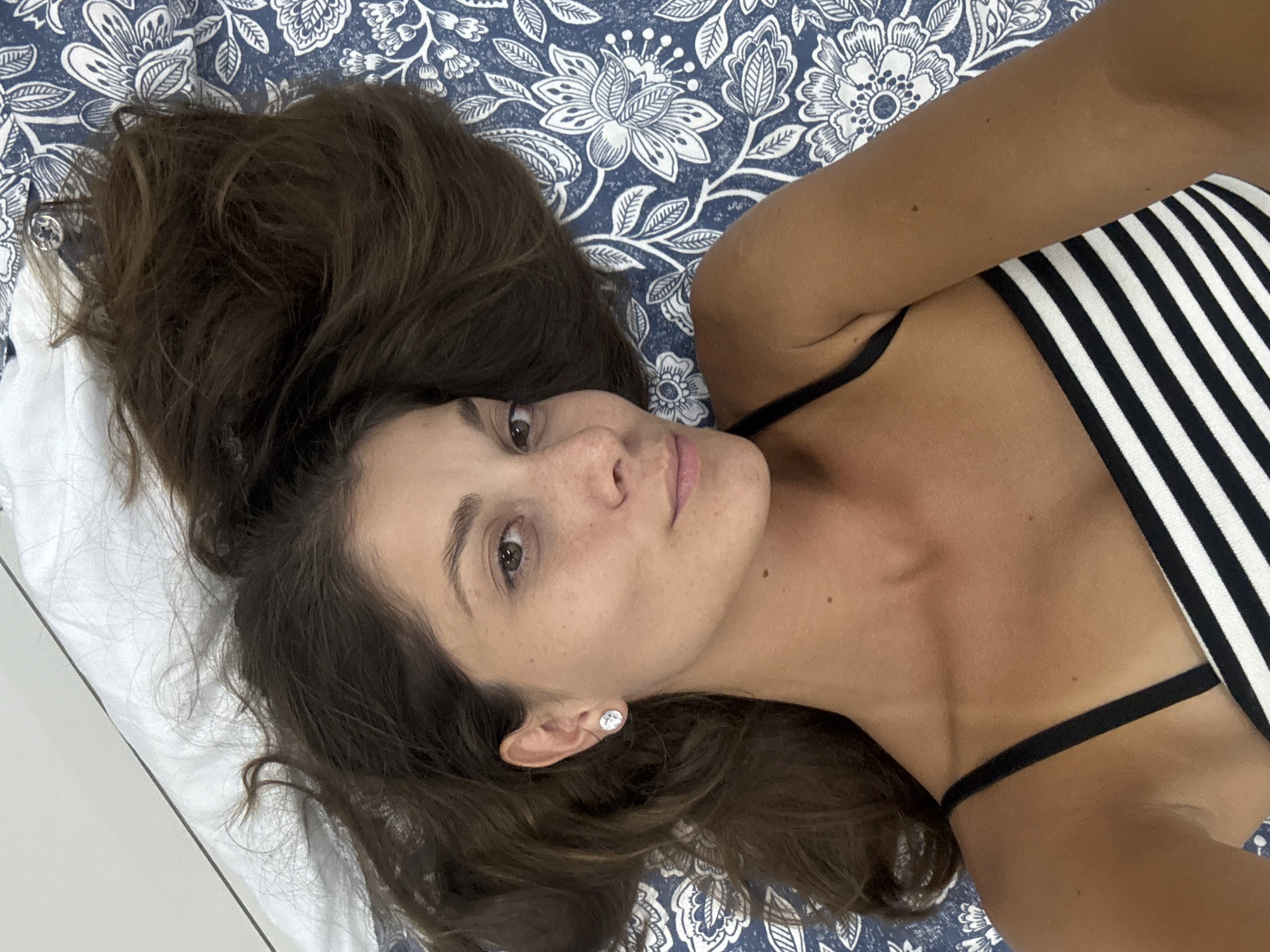 A woman with long brown hair is lying on a bed with a blue and white patterned bedspread. She is wearing a black and white striped top with black straps. She has an earring in her left ear. Her left arm is raised above her head. The image was not taken in Kuala Lumpur, Malaysia, but most likely in a private bedroom, based on the bed and surroundings.