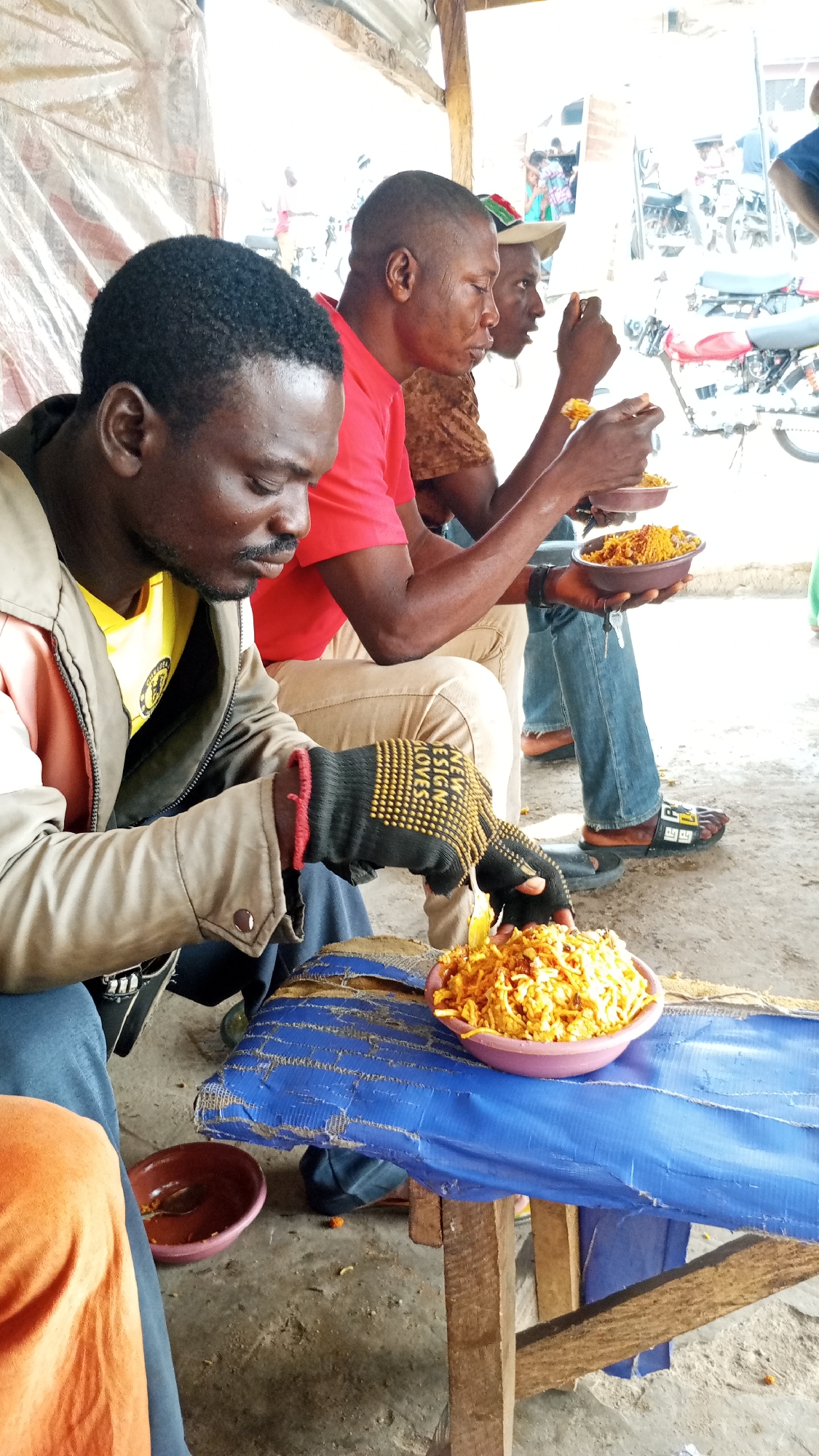 In Adankolo, Nigeria, three African men sit side-by-side at a makeshift table, eating a meal of what appears to be noodles from small bowls. The man closest to the camera wears a yellow shirt, a tan jacket, and black gloves with yellow dots. He is reaching into a large bowl of noodles on the table. The table is made of wood and covered with blue material. The other two men, one in a red shirt and the other in a brown shirt and baseball cap, are actively eating from their bowls. In the background, there are motorcycles and other people. The scene appears to be outdoors under a sheltered area.