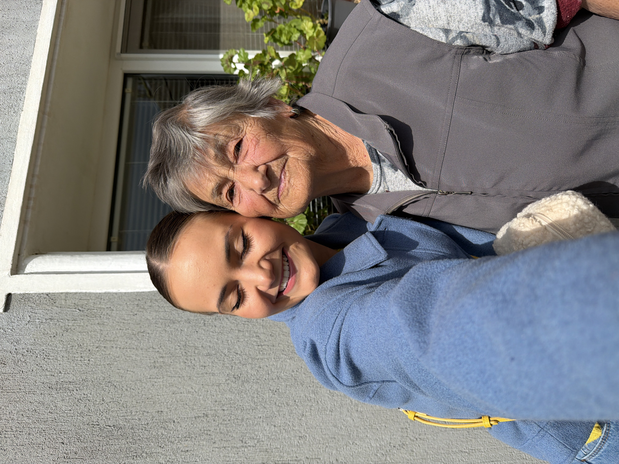 An elderly woman and a younger woman pose closely together outdoors. The older woman has gray hair and is wearing a gray vest over a gray patterned top. She is smiling with her head resting on the younger woman. The younger woman has dark hair pulled back, and is wearing a blue jacket. She is also smiling. The background shows part of a building with a window, a gray wall, and some green foliage. The location is outdoors in bright sunlight, but information to specifically identify it as being in the city of Sofia, Bulgaria is not present.