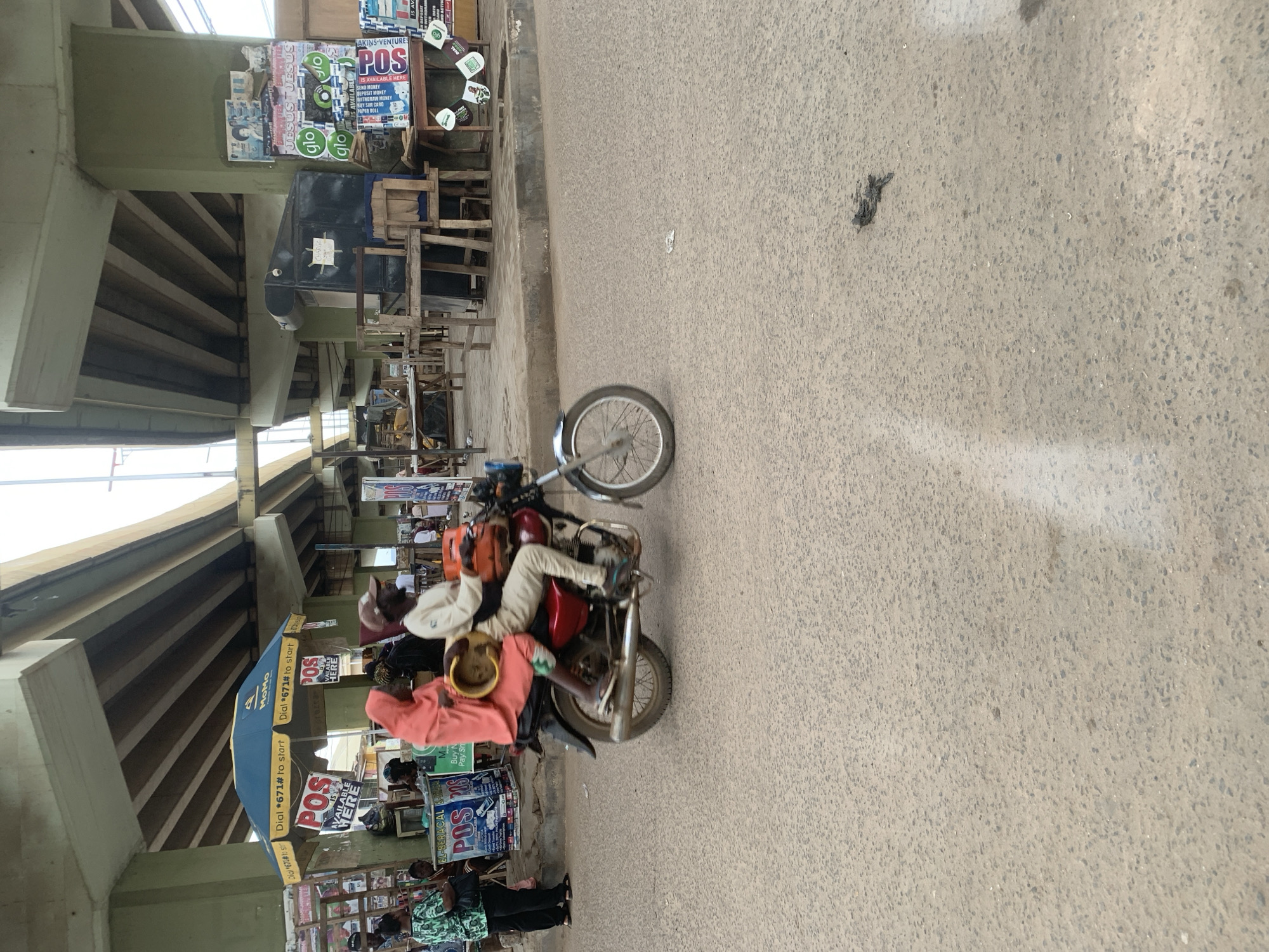 In Shagamu, Nigeria, under an elevated road structure, a man and a woman are riding on a red motorcycle. The man, wearing a light-colored shirt and pants, drives the motorcycle. The woman, dressed in a pink hijab and robe, sits behind him and holds a yellow container. They are moving from left to right across a paved road, which appears to be a street in a busy urban area. To the left of the road, there are several stalls and umbrellas, some displaying advertisements for "POS" services. A person in a green and white patterned shirt stands near these stalls. The environment is consistent with a market or commercial area in a Nigerian city, characterized by a mix of street vendors, traffic, and pedestrian activity.
