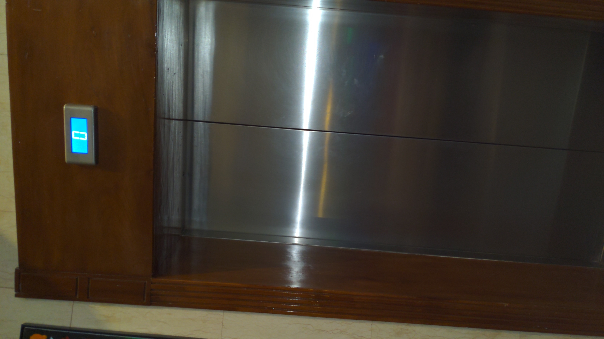 A close-up shot shows a section of a wooden panel with a metallic elevator door embedded within it. A blue-lit elevator call button is visible on the left side of the wooden panel. The location is not discernible from this image.