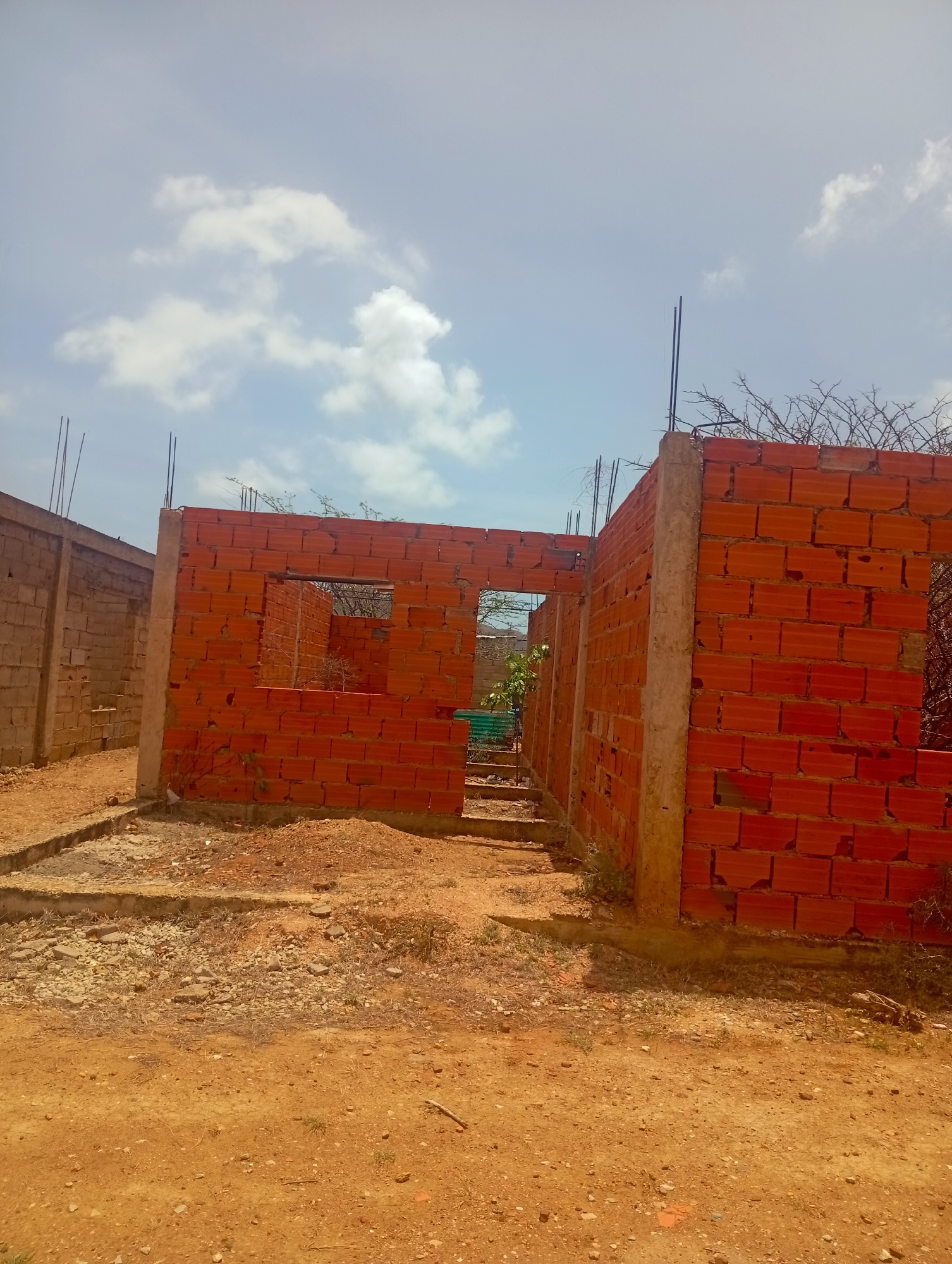 In Altagracia, Venezuela, a partially constructed building made of red bricks and concrete columns stands under a bright, cloudy sky. The structure has open doorways and windows, revealing dried vegetation within. Rebar protrudes from the tops of the walls, indicating ongoing construction. The ground is covered in dirt, gravel, and some sparse dry grass. To the left, another unfinished building constructed from grey blocks is visible. The overall scene depicts an active construction site in a dry, possibly arid, environment.