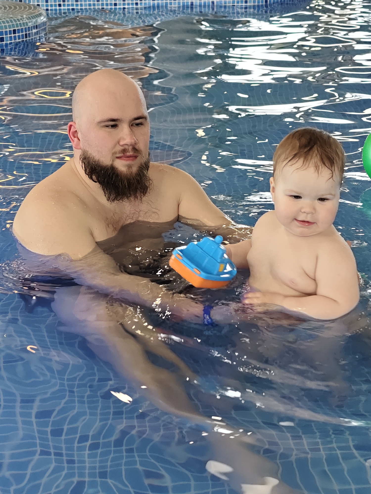 In an indoor swimming pool in Votkinsk, Russia, a man and a young child are engaged in water play. The man, positioned on the left side of the frame, has a bald head, a full dark beard, and a mustache. He is shirtless and submerged in the blue water up to his chest, looking slightly downwards and to his right with a neutral, perhaps thoughtful, expression.
To the man's right, a baby or toddler with light brown hair sits upright in the water, also shirtless and submerged to its chest. The child has a gentle smile and looks towards its right, appearing content. In the child's left hand, or floating nearby, is a bright blue and orange toy boat. A portion of a green object, possibly another toy or flotation device, is visible at the far right edge of the image.
The setting is clearly an indoor swimming pool, with light blue mosaic tiles covering the pool's bottom and visible along the edge. The water is calm and clear, reflecting light on its surface. The scene suggests a pleasant and supervised activity, possibly a swimming lesson or recreational time, for the child. No text is visible in the image.
