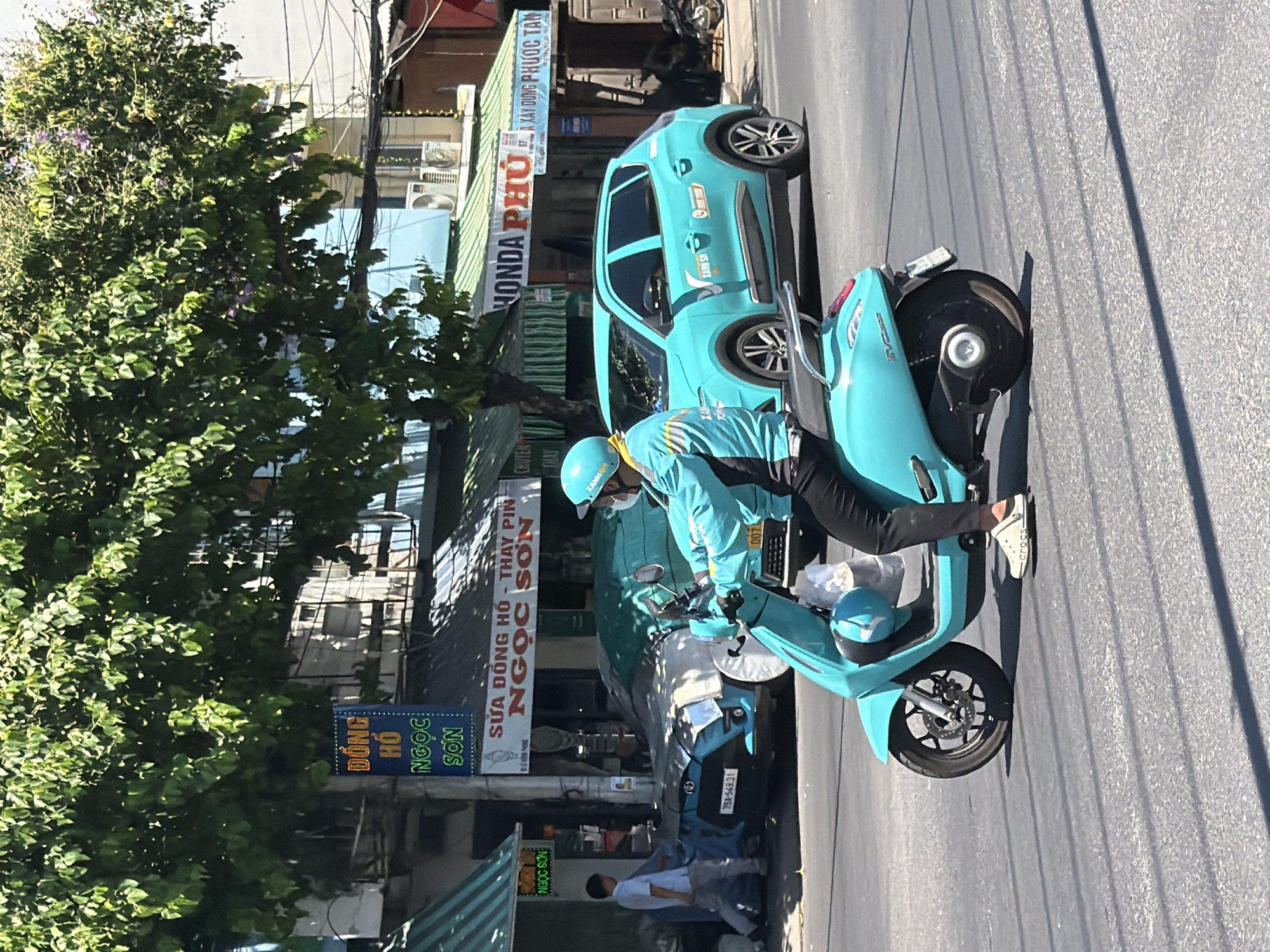 A person wearing a blue jacket, white mask, and blue helmet is riding a teal scooter on a paved street in Nha Trang Ward, Vietnam. The scooter has a white helmet resting on its front. In the background, a teal car is parked on the side of the road. Behind the car, buildings with green awnings are visible, displaying signs with Vietnamese text. One sign reads "NGỌC SƠN," and another features "HONDA PHÚ." A person in a white shirt and grey pants is walking towards the buildings on the left. The scene is illuminated by sunlight, casting shadows on the street. The environment appears to be a commercial area with shops and vehicles.