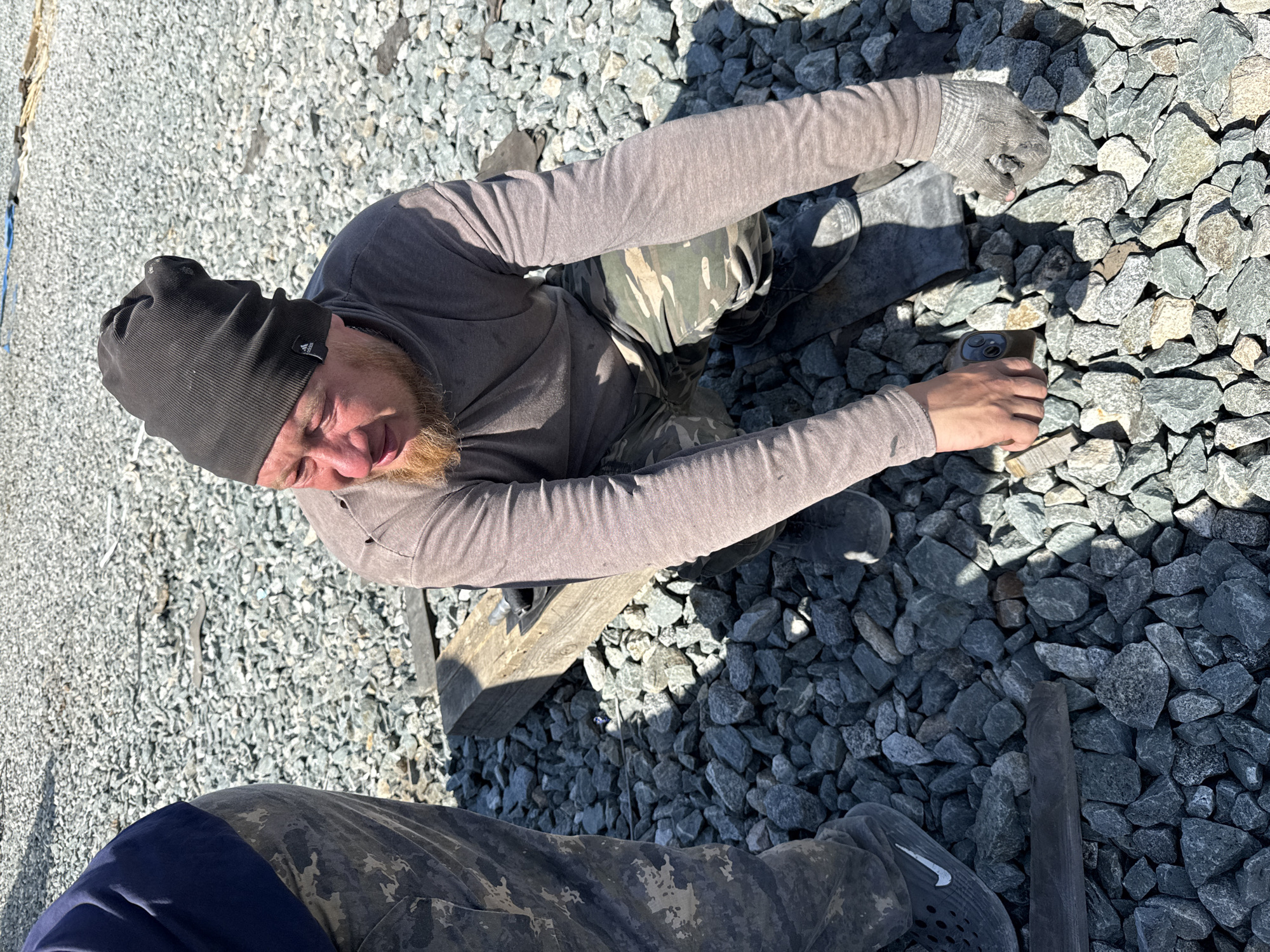 The image shows a man outdoors surrounded by gravel. The man is wearing a dark-colored beanie, a long-sleeved gray shirt, and camouflage pants. He is kneeling or sitting amidst the gravel. He is holding a smartphone with his right hand. He wears gloves on his left hand. The location cannot be confirmed to be Leninsk, Russia based on the image alone.