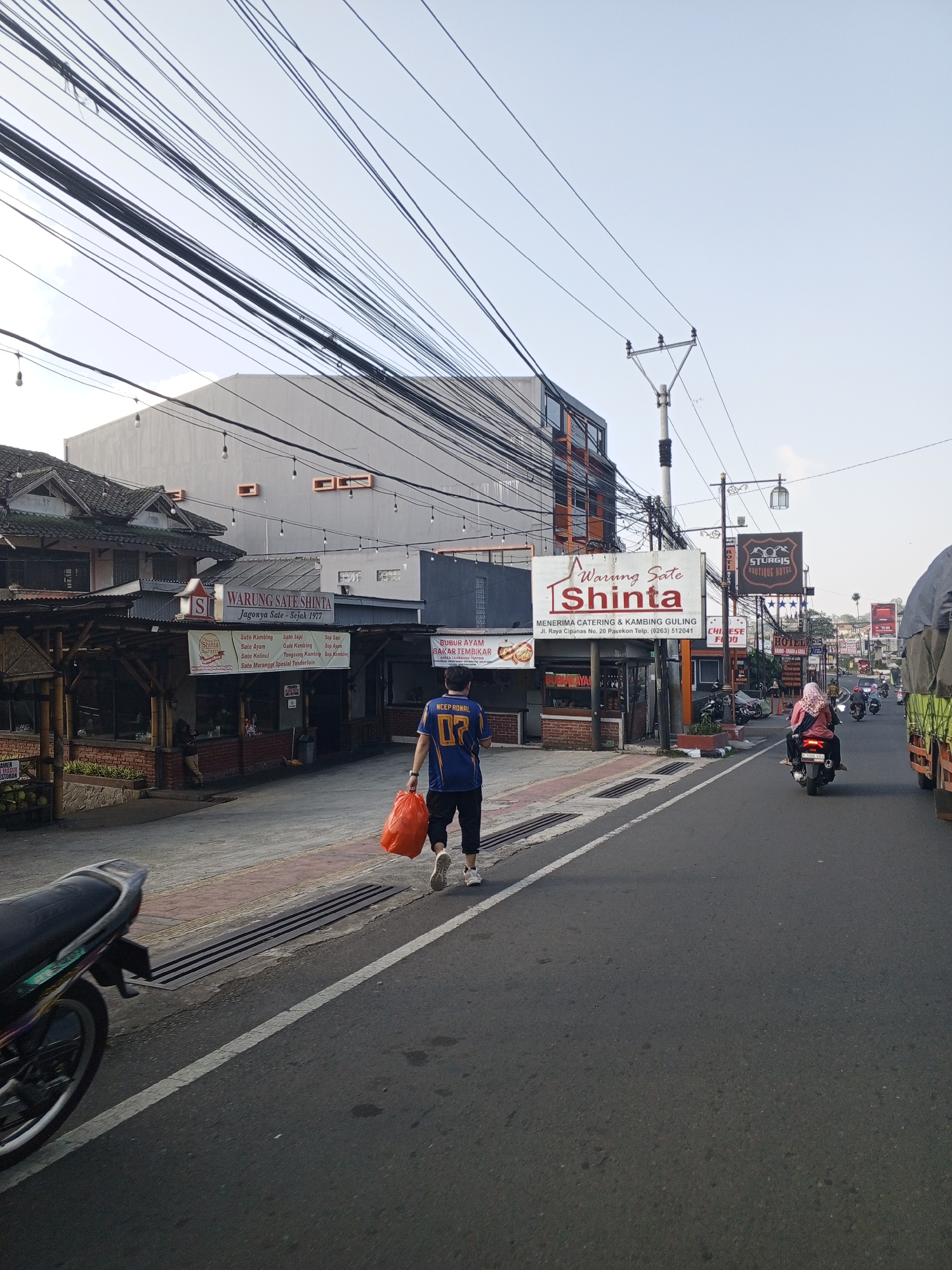 A daytime street-level image captures a commercial road scene in Cipanas, Cianjur Regency, West Java, Indonesia, as evidenced by multiple signs displaying Indonesian language and a specific street address and telephone number corresponding to that region. This location information contradicts the provided detail of Weddagala, Sri Lanka. In the foreground, the lower-left section shows the rear wheel and part of a dark-colored motorcycle, likely a scooter, parked on a paved curb. Adjacent to it, a red-brown section of the sidewalk transitions to a dark grey asphalt road, marked by a solid white line separating the shoulder from the main lane. A rectangular metal drainage grate is embedded in the curb area. Walking away from the camera on the road's shoulder is a man wearing a blue short-sleeved shirt with yellow and gold details, including "NCEP RONAL" and "07" on the back. He wears dark shorts and white athletic shoes and carries a bright orange plastic bag in his left hand. The midground features "Warung Sate Shinta," a restaurant complex on the left side of the road. It has a dark tiled roof, open-air seating areas with bamboo and wooden post structures, and a prominent white sign detailing its menu items such as "Sate Kambing," "Sate Ayam," and "Sate Maranggi Spesial Tenderloin." The sign also advertises "MENERIMA CATERING & KAMBING GULING" and provides the address "Jl. Raya Cipanas No. 20 Pasekon Telp. (0263) 512041." String lights with bare bulbs hang across the restaurant's facade. A solitary figure in dark clothing is seated at one of the restaurant's tables. Behind the restaurant, a multi-story grey building with horizontal orange accents and dark-framed windows is visible. A dense network of black utility cables crisscrosses the sky directly above and in front of these buildings. On the right side of the road, a grey metal utility pole with insulators stands prominently. Further down the road, a larger white sign also for "Warung Sate Shinta" is seen, followed by signs for "Sturgis Boutique Hotel," a generic "HOTEL" sign with "BAR & GRILL," and a partially visible "CHINESE FOOD" sign. Several vehicles, predominantly motorcycles, are traveling down the road. A woman wearing a pink hijab and dark clothing rides a black scooter away from the camera. The far right edge of the frame shows the front portion of a large truck with an olive-green tarp covering its load. The background consists of additional commercial buildings, utility