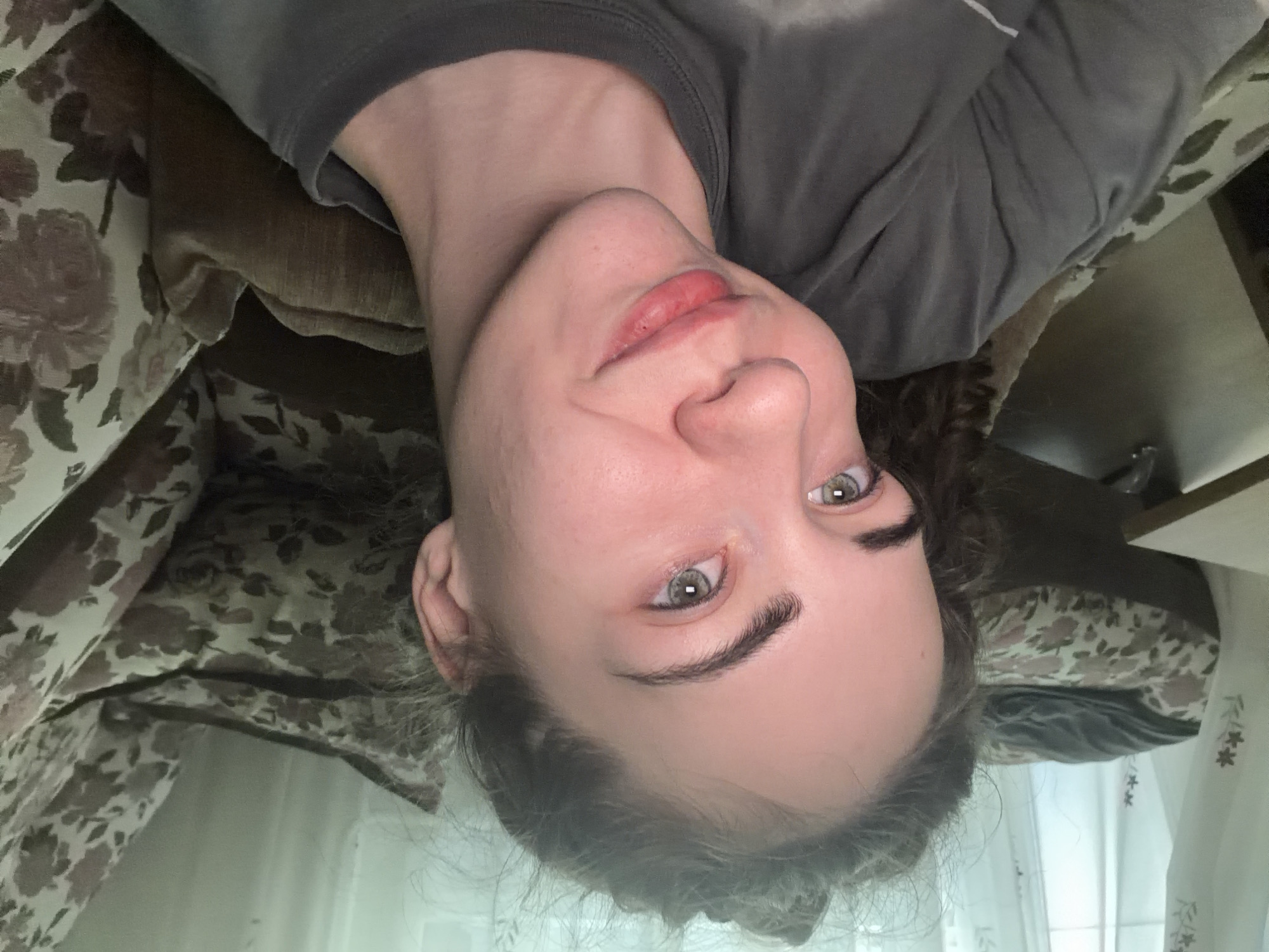 A young woman with light skin, light brown hair pulled back in a bun, and green eyes is pictured in a close-up selfie. She is wearing a grey t-shirt. The background shows a couch with a floral pattern and a window with sheer curtains. The lighting is soft and diffused. The woman is smiling slightly, and her head is tilted. There is no discernible information within the image to determine her location in the city of Zavodoukovsk, Russia.