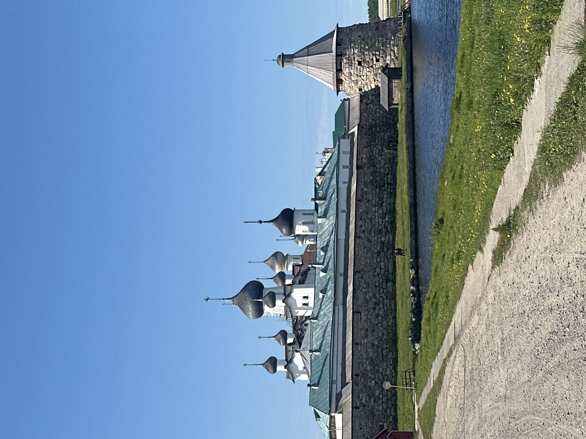 The image depicts the Solovetsky Monastery in Solovetskiy, Russia, under a clear blue sky. A stone wall, topped with a wooden structure, encloses the monastery complex which features multiple white buildings with dark, onion-shaped domes and copper roofs. A circular stone tower with a conical wooden roof stands to the right of the monastery, adjacent to a small wooden shed. In the foreground, a gravel path leads towards the monastery, with a grassy area dotted with yellow wildflowers bordering a body of water. Two figures, appearing to be people, are visible on the grass near the water's edge. A small red building is partially visible on the far left of the image.