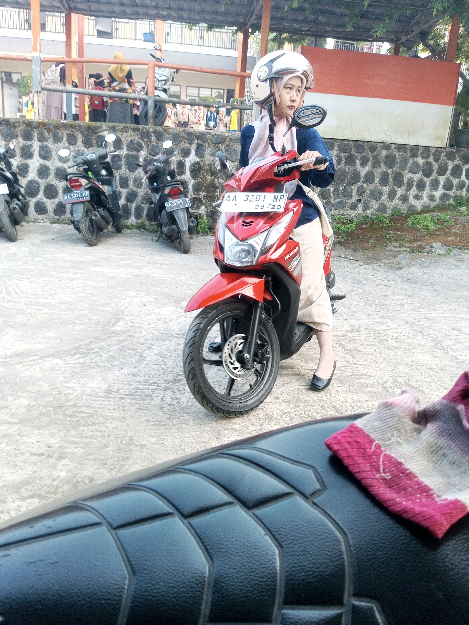 A woman in a white helmet and a blue and pink outfit is sitting on a red scooter in what appears to be a parking lot in Krasak, Indonesia. The scooter has a license plate that reads "AA 3201 NP 09-29". Behind her, there is a stone wall and a building with a red and white banner. Several other scooters are parked in the background, and some people, possibly students, are visible near the building. The ground is paved with concrete. A fabric with pink and white stripes is resting on the seat of another scooter in the foreground.