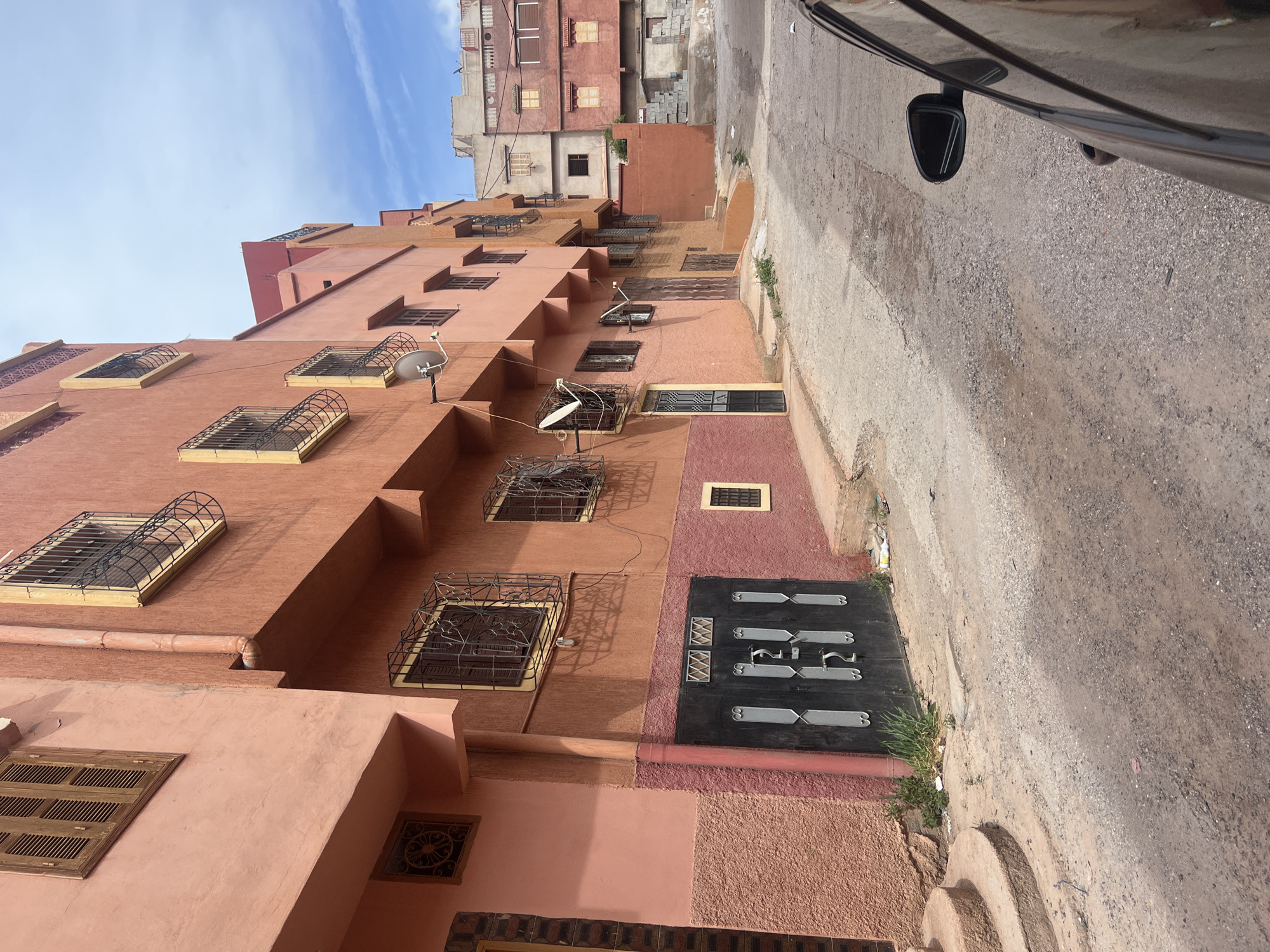 The image shows a narrow street in Ouaouizert, Morocco, with a multi-story building on the left side and a road on the right. The building is composed of different shades of orange and features windows with decorative metal grilles and satellite dishes attached to the facade. The door is dark with silver-colored decorative elements. The road shows signs of wear and tear. The sky is visible in the background. A car is visible on the top right corner of the photo.