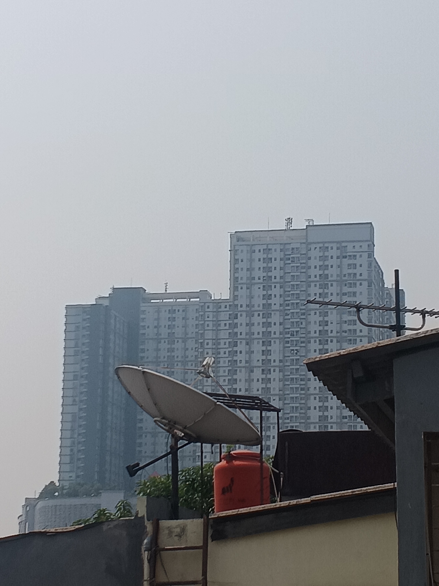 The image captures a view of high-rise apartment buildings in a hazy urban environment, likely West Jakarta, Indonesia. In the foreground, a satellite dish is prominently positioned, supported by a metal structure. Adjacent to the dish, an orange water tank rests on a rooftop. Further back, another large apartment building complex is visible, its facade covered in numerous small windows. A portion of a rooftop with dark roofing material and a sloped overhang is also present in the immediate foreground. The sky is overcast and hazy, obscuring distant details and contributing to a muted color palette. The overall scene depicts a densely populated urban landscape with residential architecture and essential infrastructure for urban living.