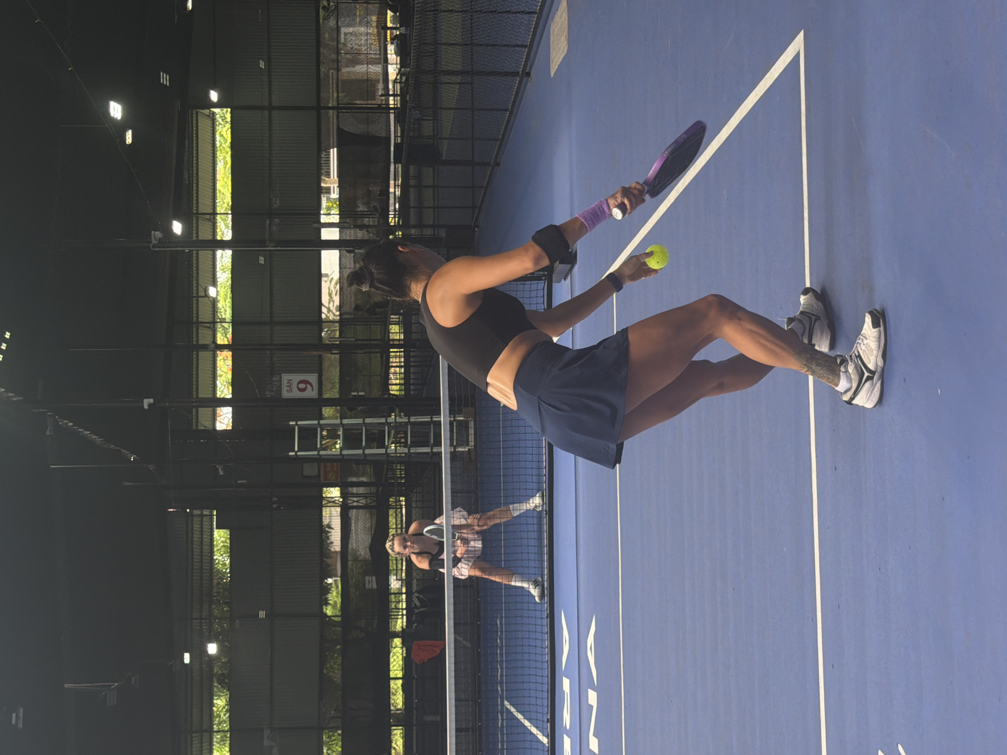 This image captures a padel or tennis match taking place on a blue court with white lines, likely in an indoor or covered facility. In the foreground, a woman stands with her back partially to the camera, holding a purple padel racket in her right hand and a yellow ball in her left, appearing ready to serve or hit. She wears a black sports bra, a dark blue skirt, white athletic shoes with dark accents, and has a purple wristband on her left arm and a dark wristband on her right. A tattoo is visible on her right calf. Her hair is tied up.
Across the net, another woman in a light-colored top and skirt is in a ready position, also holding a padel racket. The court is illuminated by a mix of natural light filtering through openings along the sides of the structure, revealing green foliage, and artificial lighting fixtures overhead. The background features metal support beams and fencing typical of a sports complex. A red and white sign with the text "SAN 9" is visible in the upper background, indicating a specific court number. The overall atmosphere suggests an active sports environment during what appears to be daytime.