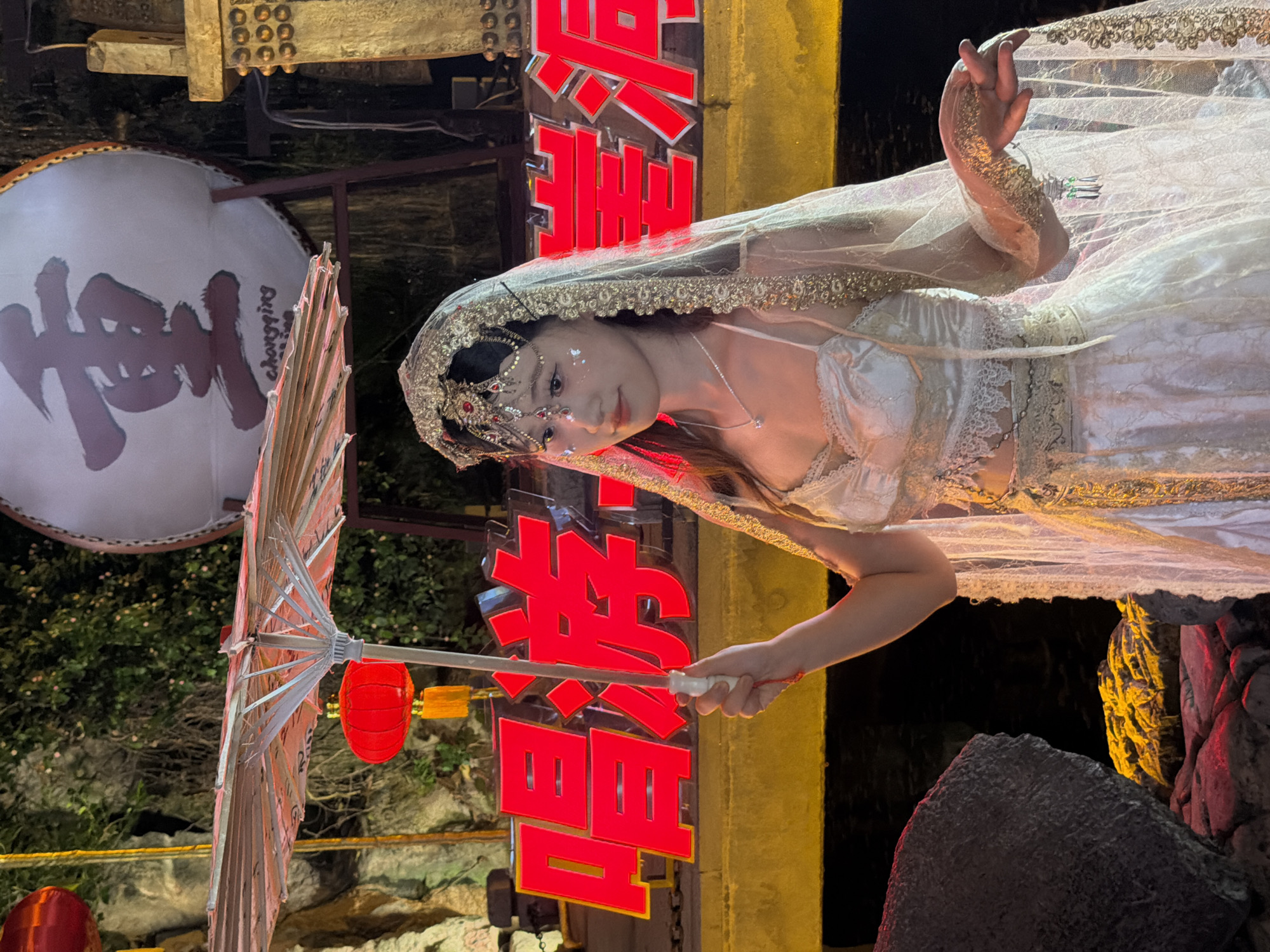 In Yuzhong District, Chongqing, China, a young woman, adorned in traditional-inspired attire and elaborate headwear with decorative sequins and jewels, stands holding a partially opened, ornate paper umbrella. Her dress, a flowing white gown with intricate lace and gold trim, complements her headdress. She is illuminated by the warm glow of neon red signage in the background, which displays Chinese characters and the word "chongqing" in English beneath a large, round drum bearing the Chinese character "重" (zhòng, meaning heavy or important). Red lanterns hang to the left, casting a soft light. The scene appears to be outdoors at night, with a dark body of water visible in the foreground and rocky formations.