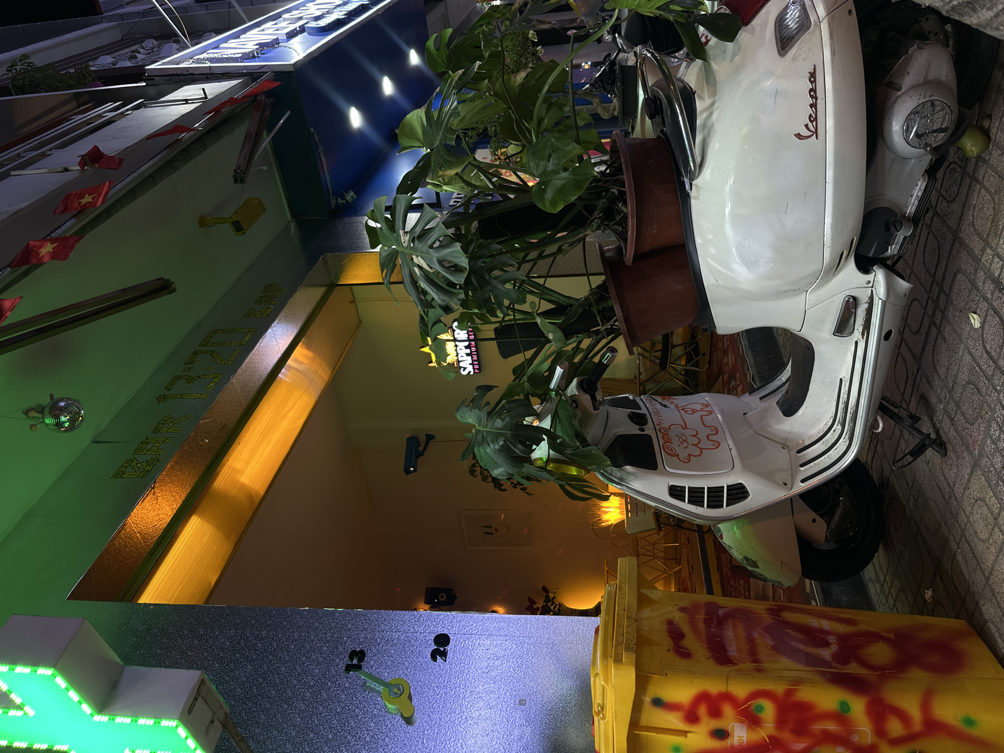 This media file captures a vibrant street scene at night, likely in Nha Trang Ward, Vietnam, given the cultural cues. The primary subject is a white vintage Vespa scooter, parked on a paved sidewalk. Uniquely, the scooter is decorated with large leafy plants, possibly Monstera, held in brown pots attached to its front, blending nature with urban transport.
Adjacent to the scooter is a bright yellow trash can, heavily covered in red graffiti, with some green and blue accents. The scooter itself has the "Vespa" logo in red script on its side and some stylized red drawings or stickers on its front mudguard.
The background features a building facade, brightly illuminated with various artificial lights, suggesting a bar or entertainment establishment. Small red flags with a yellow star, characteristic of the Vietnamese national flag, hang vertically along the building's exterior. Above these flags, a security camera is visible.
Several signs are present: a large blue sign with illuminated white letters, where "BAR" and "320" are partially discernible, and another sign further down showing "SAPPORO PREMIUM BEER." A green neon cross-shaped sign and illuminated numbers "18" and "20" also adorn the building. A small disco ball hangs from an awning, reflecting light. Warm yellow light emanates from inside the establishment, contrasting with cooler blue and green lights elsewhere. No specific activity or people are depicted; the scene is static, capturing the eclectic details of a bustling city street after dark.