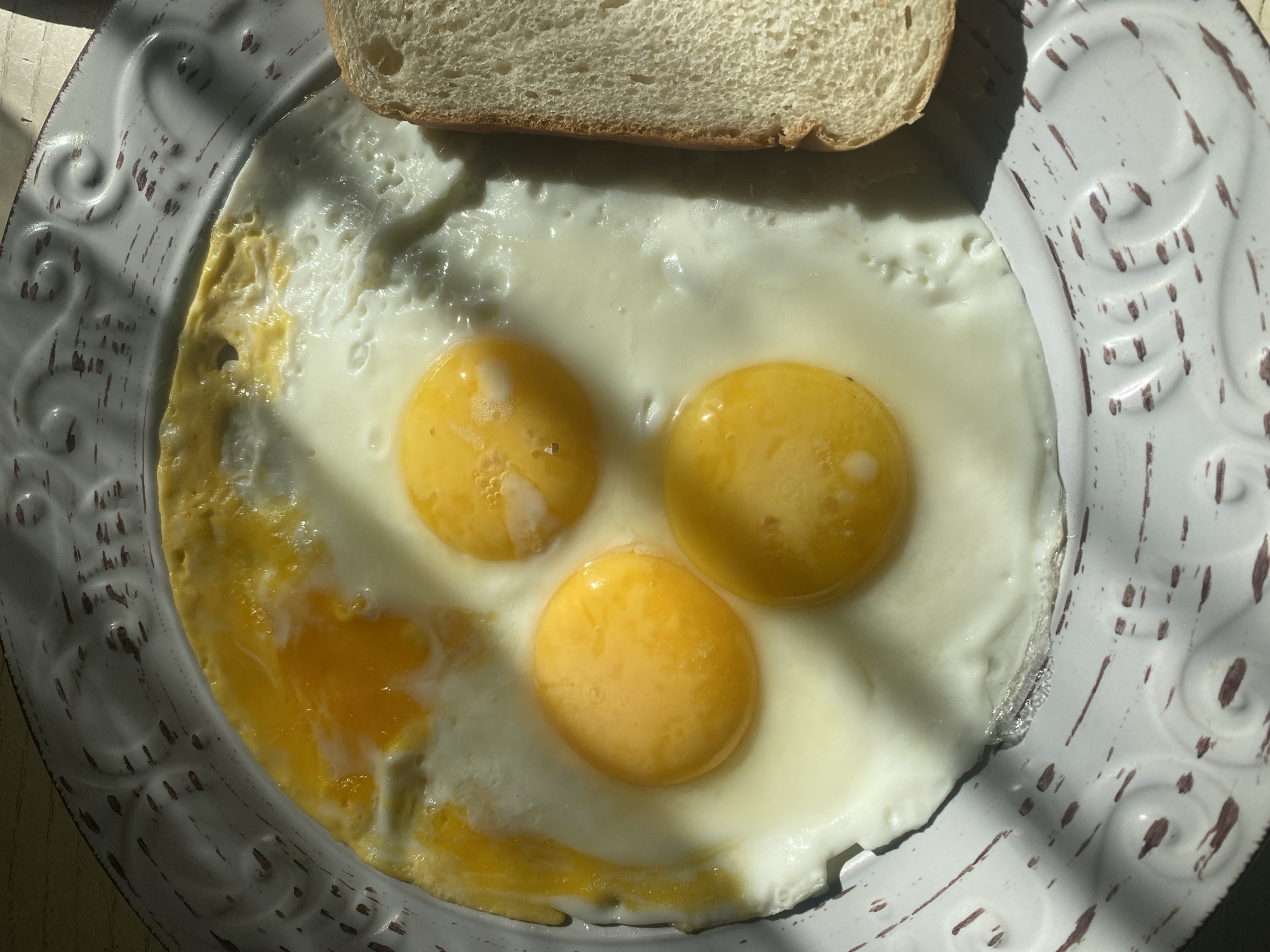 An overhead, close-up shot shows three fried eggs and a slice of white bread on a white, embossed plate. The eggs are sunny-side up, with bright yellow yolks. The whites are cooked through and slightly bubbly in places. The bread is toasted on one side and appears soft on the other. The plate has a rustic, distressed finish with dark brown marks on the white surface. The scene is lit by natural sunlight, casting shadows across the plate. The location is not specified in the image or its metadata.