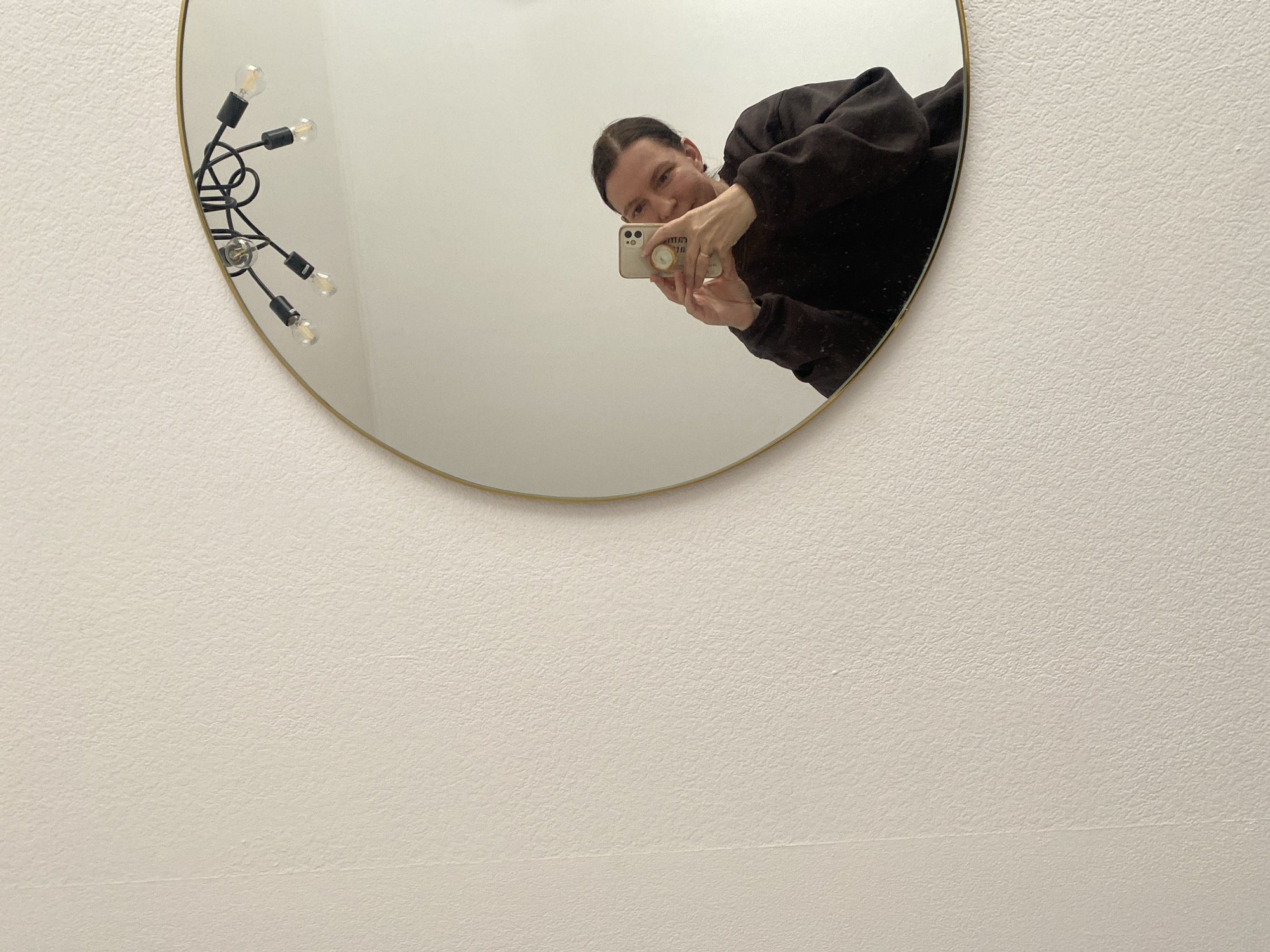 A person is taking a selfie in a round mirror. The mirror is mounted on a white wall and has a gold trim. To the left of the person's reflection, a light fixture with multiple exposed bulbs is visible on the mirror. The person is wearing a dark-colored sweatshirt and holding a smartphone. The image doesn't provide information about the location in Yekaterinburg, Russia.