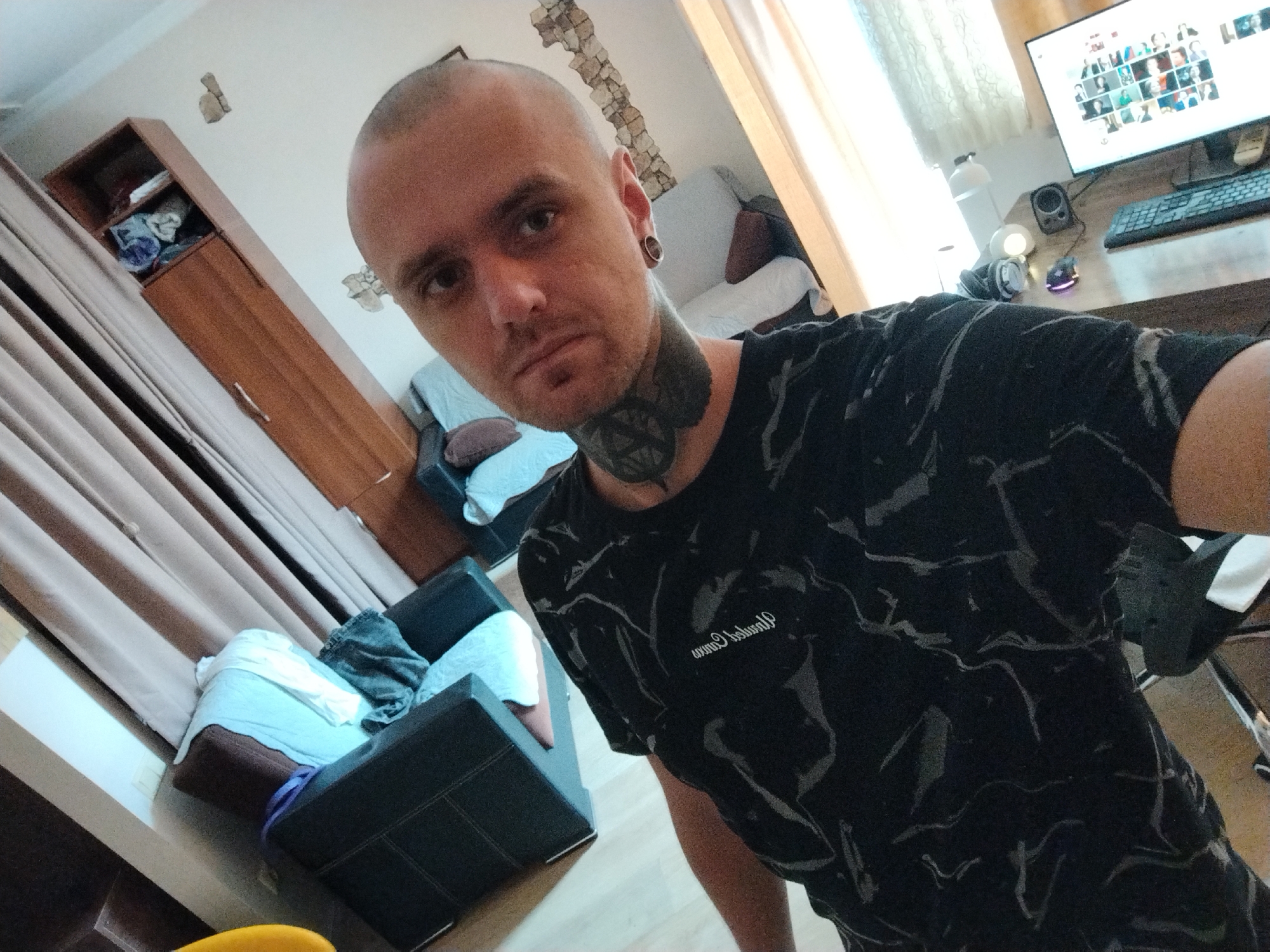 The image shows a man with a shaved head and tattoos on his neck. He is wearing a black t-shirt with a marble pattern. He is standing in a room that appears to be a living room or office. There is a computer monitor on a desk in the background, as well as a couch and a wardrobe. The room is lit by natural light coming from a window. The image is taken from a high angle, looking down at the man. The man is looking directly at the camera. The overall impression is that this is a selfie taken in a home environment.