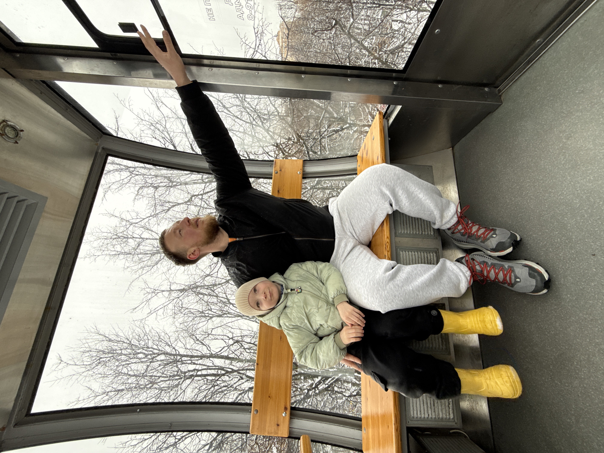 Inside a viewing booth, likely at the observation point of the "Permyak" monument in Perm, Russia, a man wearing a black jacket and grey sweatpants is seated on a wooden bench, with a child in a light green jacket, black pants, and yellow boots resting on his lap. The man reaches up to adjust a panel in the glass roof of the booth. Outside, bare trees and a grey sky are visible. The floor of the booth is grey.