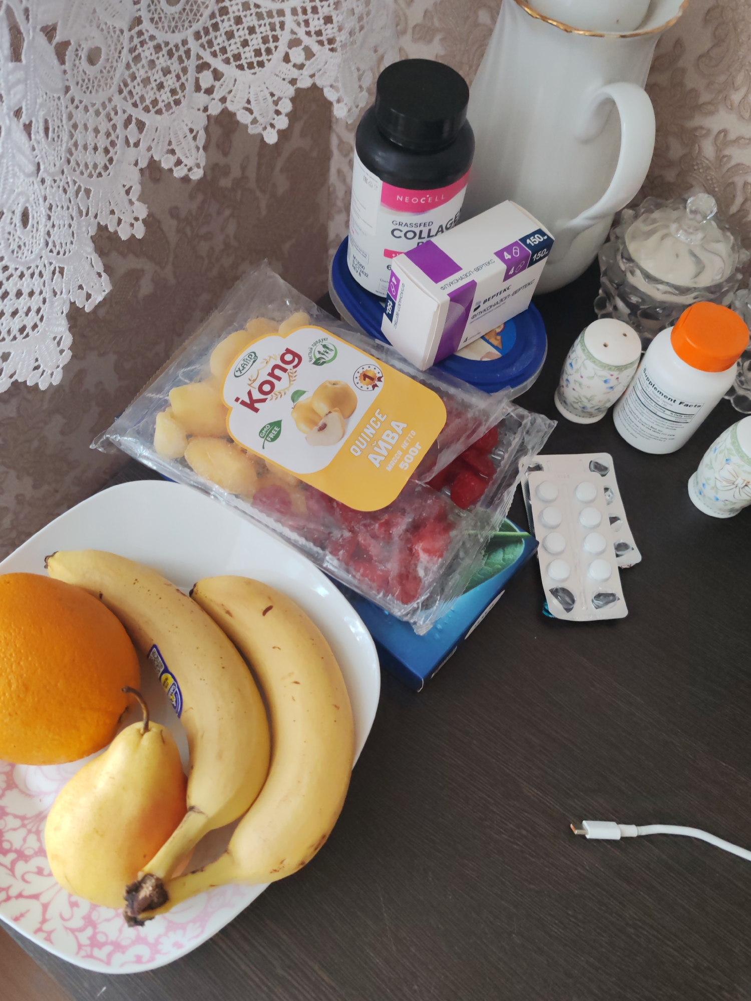The image depicts a cluttered tabletop scene with a mix of food items and various objects. The scene includes:
* A white plate containing an orange, two bananas, and a pear.
* A package labeled "Kong Quince Ayva" containing both quince pieces and red berries.
* A container of Neocell Grassfed Collagen, a white box labeled "Fluconazole-Verteks", an orange supplement bottle, and a salt and pepper shaker.
* A blister pack of white pills with some pills already removed.
* A charger cable.
* A white pitcher.
The location information (Petropavlovsk-Kamchatsky, Russia) is not discernible from the image and can't be confirmed. No people are visible, and there are no visible actions. The environment is a dark brown surface with a white, lace-like curtain partially visible in the background.