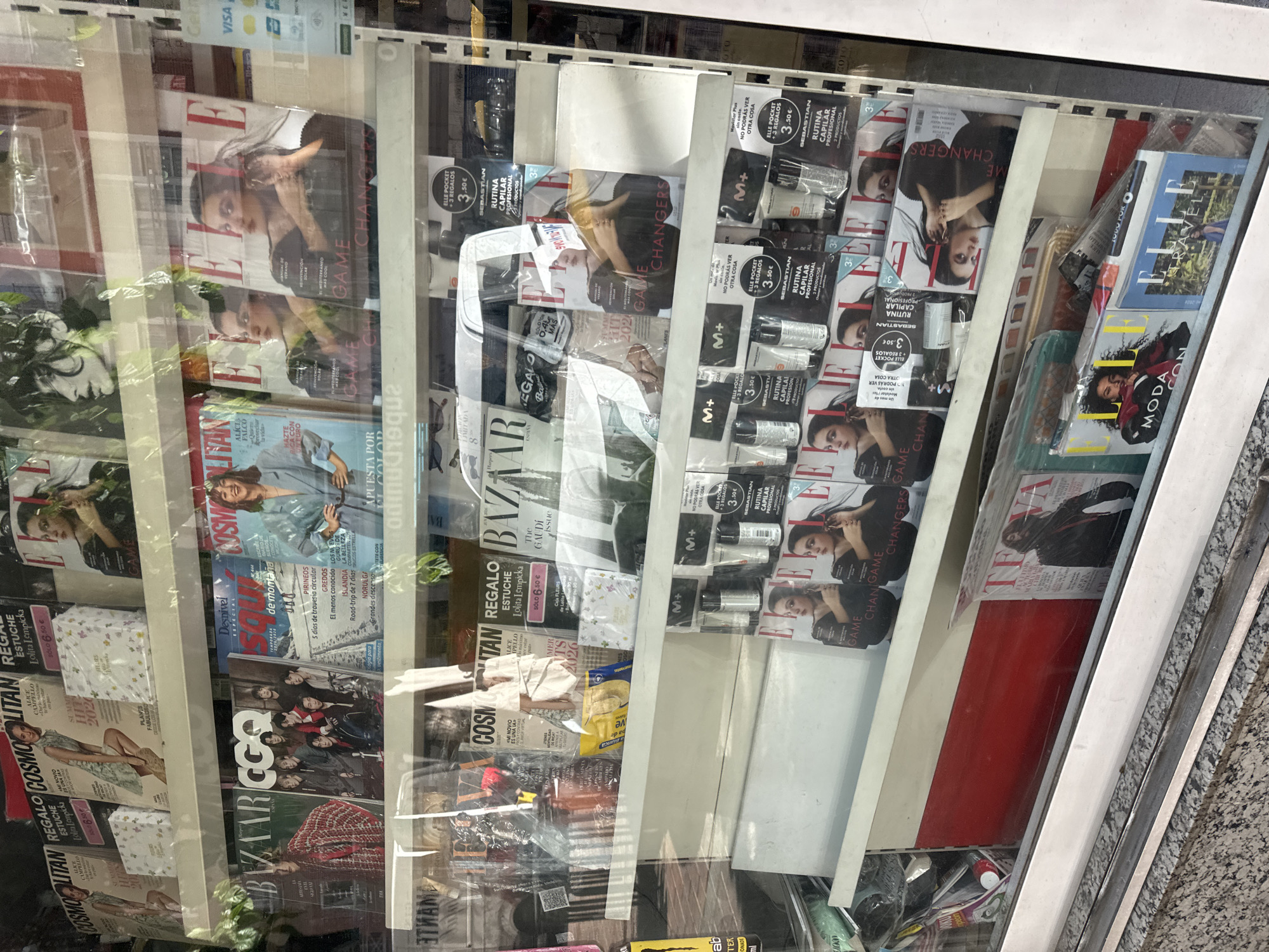 This is a medium shot of a newspaper and magazine stand, seen through a glass window. The stand is filled with various publications, many of which are fashion magazines such as Vogue, Elle, and Cosmopolitan. There are also lifestyle magazines like GQ and Harper's Bazaar. The magazines are neatly displayed on shelves.
On the top shelf, there are several magazines including Cosmopolitan with a woman in a green dress on the cover, and Elle magazines with women on the covers. Some magazines appear to have gift promotions attached to them.
The middle shelf features a wider variety of magazines. A prominent magazine is Harper's Bazaar with a woman in a red and black patterned jacket on the cover. Next to it are GQ magazines featuring a group of men. Desnivel and Cosmopolitan magazines are also visible.
The bottom shelves are also filled with magazines. Elle magazines with women on the covers are stacked together. Towards the bottom, a magazine titled "TEFA" is visible, featuring a woman in a black jacket. An "Elle Travel" magazine is also present.
Scattered among the magazines are various promotional items, including what appear to be small bottles of hair products or cosmetics, possibly gifts with purchase. There are also some items wrapped in plastic.
The overall environment appears to be an indoor retail space, likely a kiosk or a shopfront, with the magazines arranged on white shelves. The lighting suggests it is daytime. The reflections on the glass indicate that this is a display viewed from the exterior.
This specific display is located at a kiosk in the Plaza Circular, Murcia, Spain. The kiosk is situated at a busy street corner.