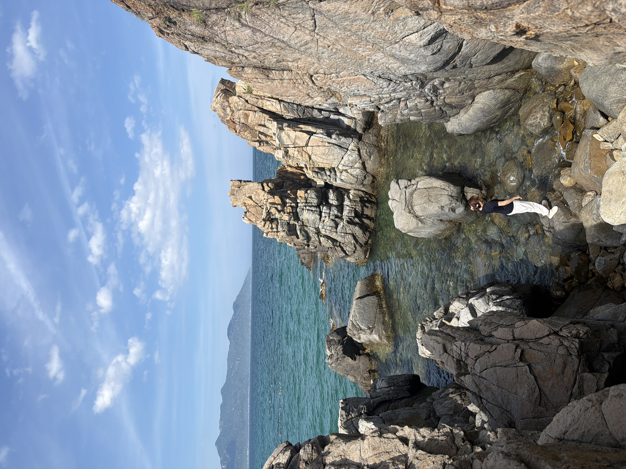 A person stands on a rocky shore in Vĩnh Lương, Vietnam, facing away from the camera. The individual is wearing a dark t-shirt and white pants. The person is positioned on a cluster of grey and tan rocks, some of which are submerged in clear, shallow water. The water is turquoise and reveals the rocky seabed beneath. Jagged, weathered rock formations rise dramatically from the water and surround the shore. In the background, the vast expanse of the sea stretches to the horizon, meeting a hazy mountain range under a bright blue sky dotted with white clouds. The scene depicts a natural coastal landscape with a solitary figure observing the environment.