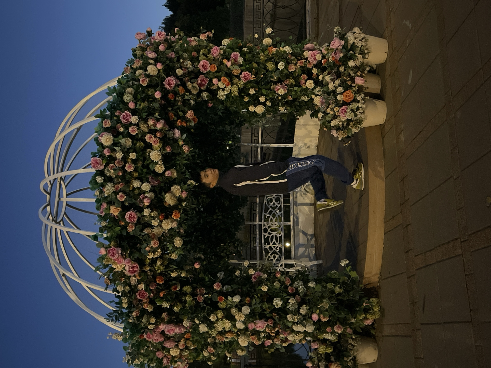 An outdoor shot captures a man in a dark blue jacket with white stripes and blue pants, with white lettering on the side, in mid-stride. He has one leg bent and is looking to his left. He is positioned inside a white, dome-shaped gazebo structure draped in an abundance of artificial pink, white, and orange flowers. The gazebo is supported by a white frame. In front of the gazebo, there are three white pots overflowing with flowers. Behind the man, a white bench with an intricate metal design is visible. The ground is paved with square tiles. The scene is set against a backdrop of a dark blue twilight sky. To the left of the gazebo, a body of water can be seen. The overall lighting suggests it is dusk or early evening.