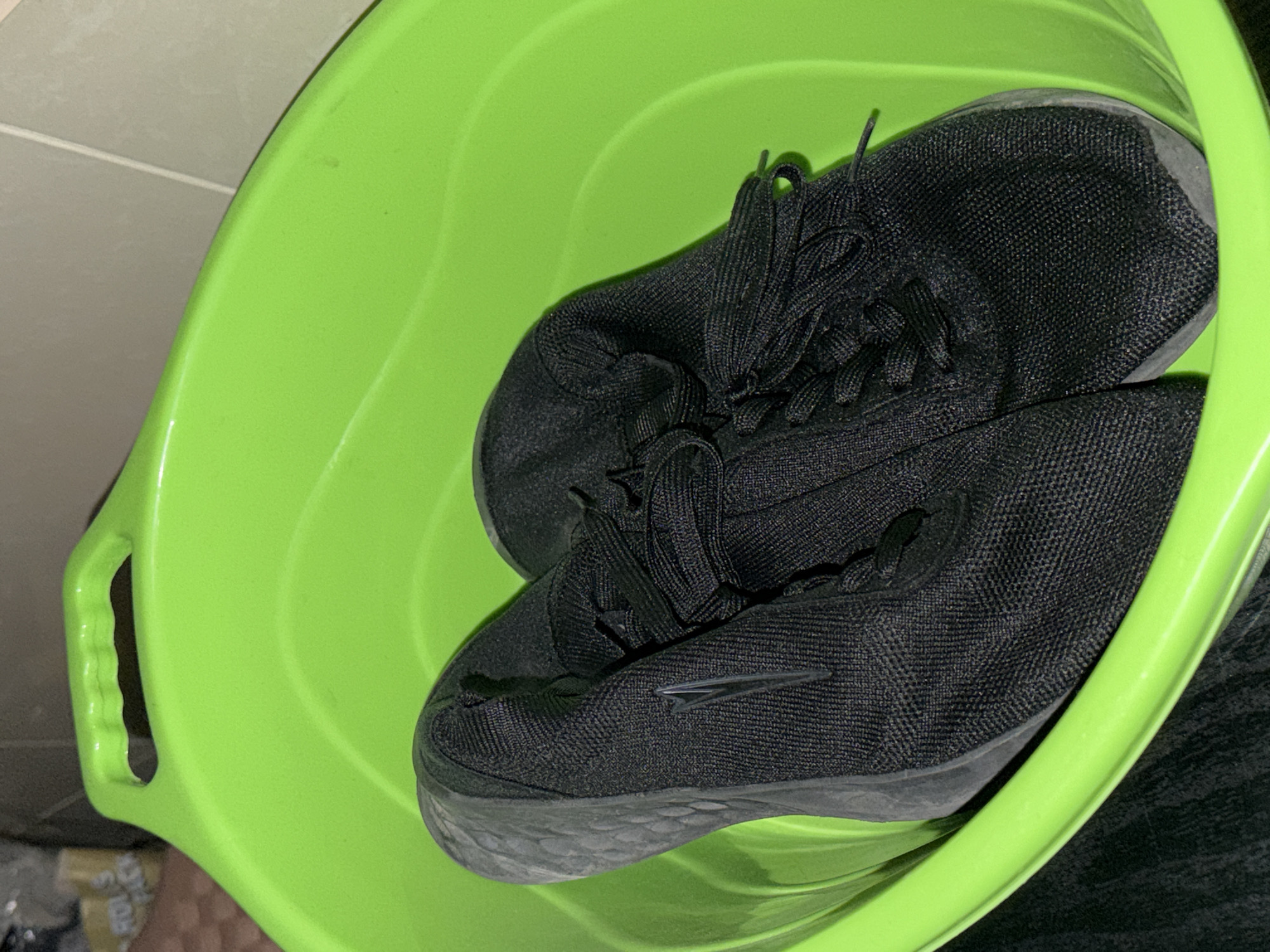 A pair of black running shoes are inside a green plastic tub. The shoes have black laces and gray soles. The background consists of a tiled wall and a dark object. The location is not identifiable as Varna, Bulgaria.