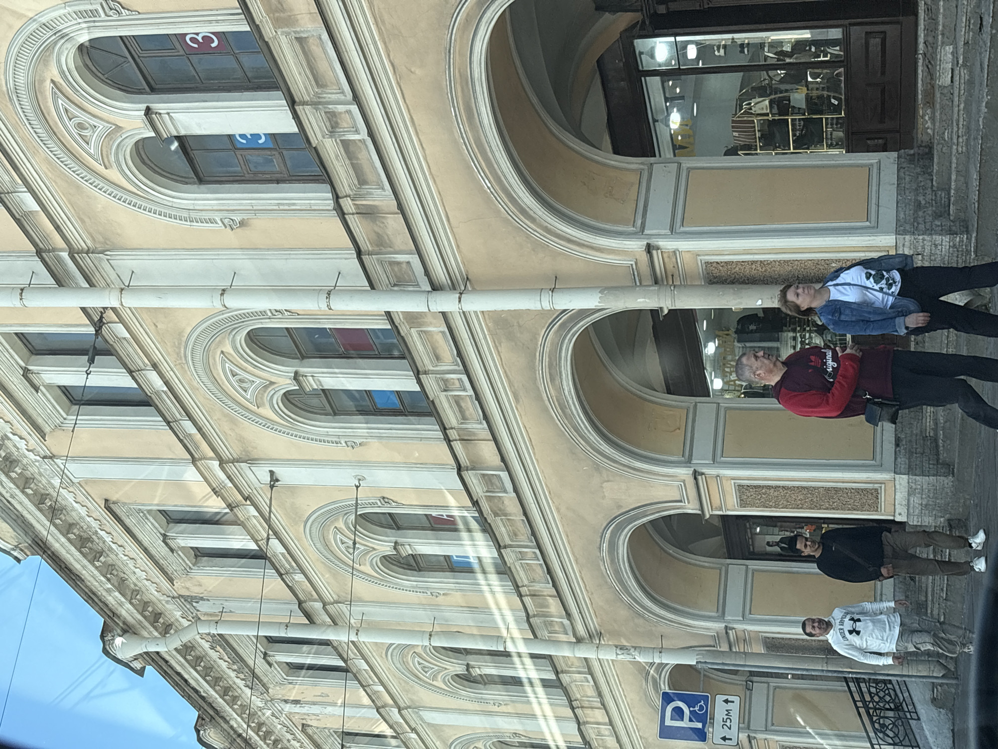 The image depicts a street scene in Saint Petersburg, Russia, focusing on the facade of a historical building with arched entrances and windows. The building is painted a pale yellow with white decorative elements. Several people are visible walking along the sidewalk. In the foreground, a man wearing a white hooded sweatshirt with "Under Armour" printed on it, and a man in a black sweatshirt are walking. Further to the right, a woman in a denim jacket and a man in a burgundy sweatshirt are also walking. The building features shops with large display windows on the ground floor. A blue parking sign with an accessibility symbol and an arrow indicating a distance of 25 meters is also present. The sky is visible at the top of the image, appearing clear and blue. Sunlight creates bright reflections on the building's facade.