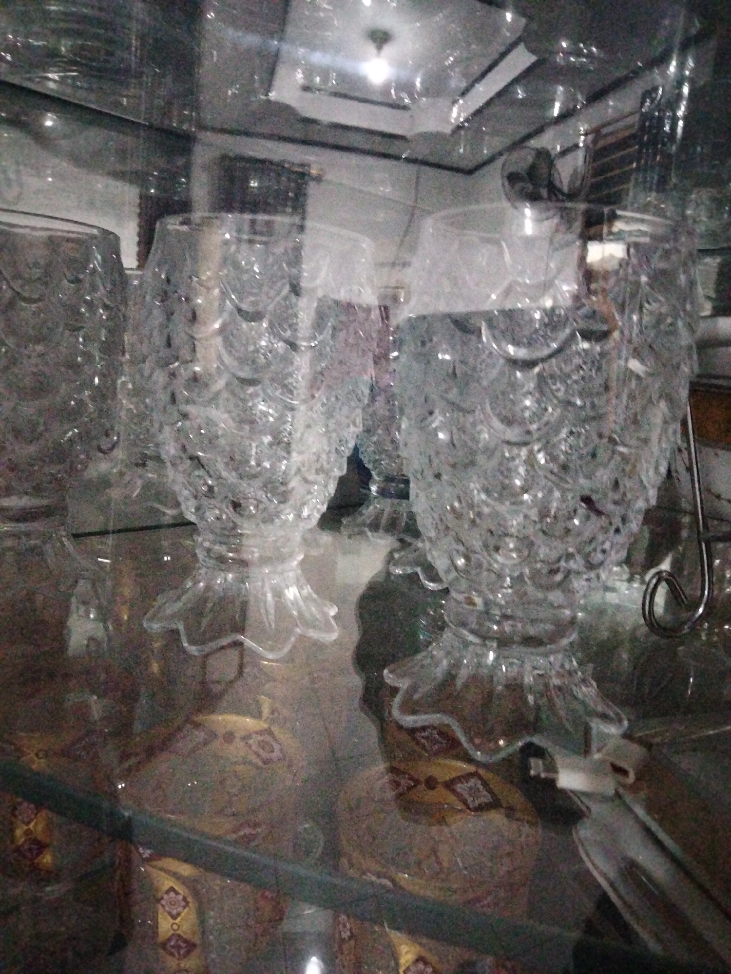 The image shows a collection of clear, intricately patterned glass objects that resemble fish. These are likely decorative vases or bowls, with a textured surface resembling scales and a fluted base. They are displayed on a glass shelf or surface, with other glass items visible in the background. The lighting is somewhat dim and appears to be artificial, with reflections on the glass surfaces.
Below the glass objects, on a lower shelf or surface, are several patterned items. These appear to be fabric-covered objects, possibly cushions or decorative elements, with a repeating geometric design in earthy tones of gold, brown, and red.
The scene is indoors, possibly a display cabinet or a shelf in a home or shop. The overall impression is one of display and decoration. There is no visible text in the image. The context of Payagaboh, Indonesia, suggests a potential cultural or traditional element to the decorative items, though this is not explicitly evident from the visual information alone. The image has a slightly grainy quality, suggesting it was taken in low light conditions with a mobile device.