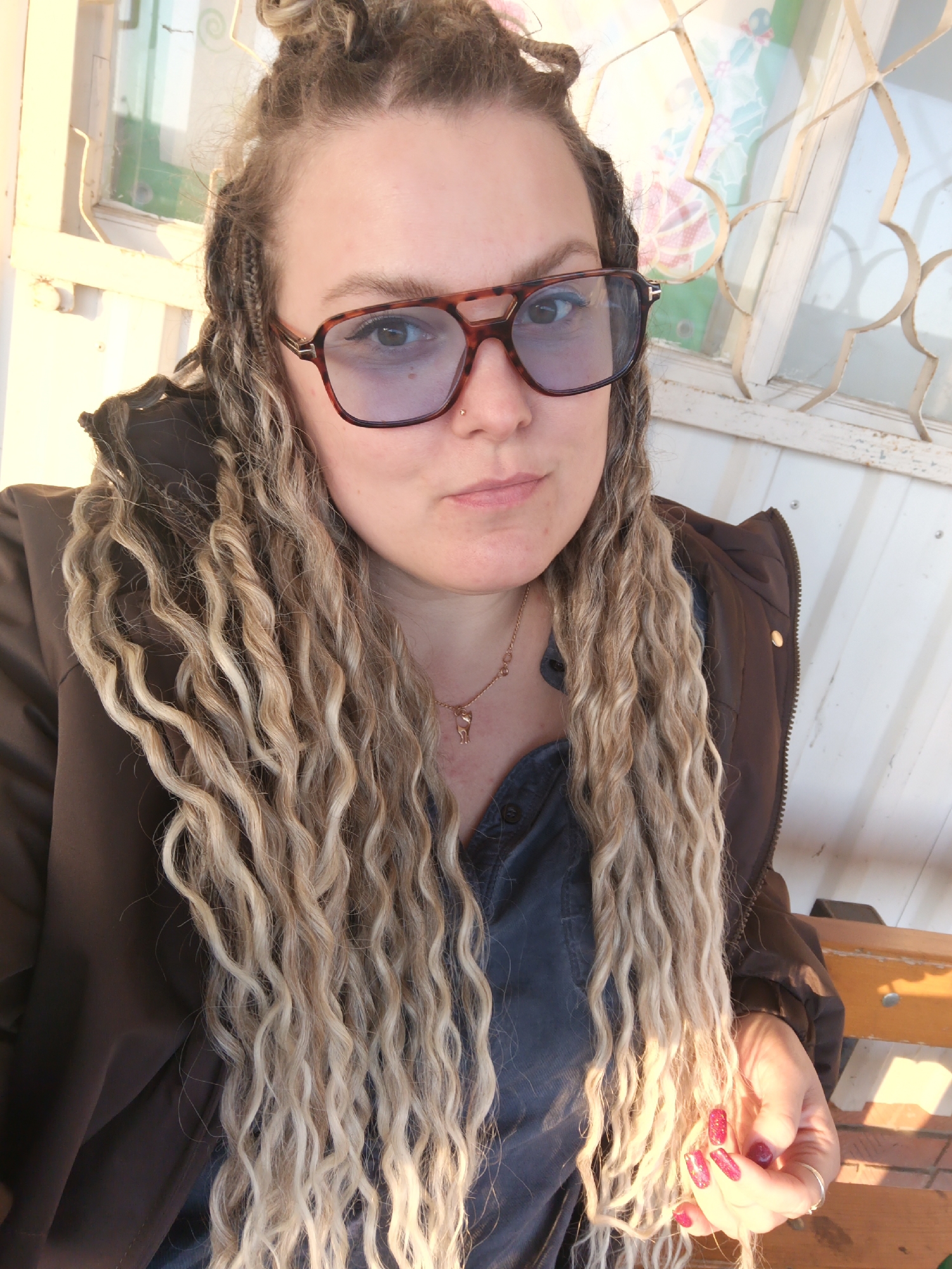 The image is a selfie of a fair-skinned woman with blonde dreadlocks, some of which are braided and tied into a bun on top of her head. She is wearing brown-rimmed glasses with blue-tinted lenses, a dark blue button-down shirt, and a brown jacket with the hood pulled up. She has a nose piercing on her left nostril and a gold necklace with a pendant. Her fingernails are painted red with glitter. The background shows a building with a window, a metal grill and a wooden bench in the foreground. The setting appears to be outdoors. This picture could have been taken in Chaikovsky, Russia.