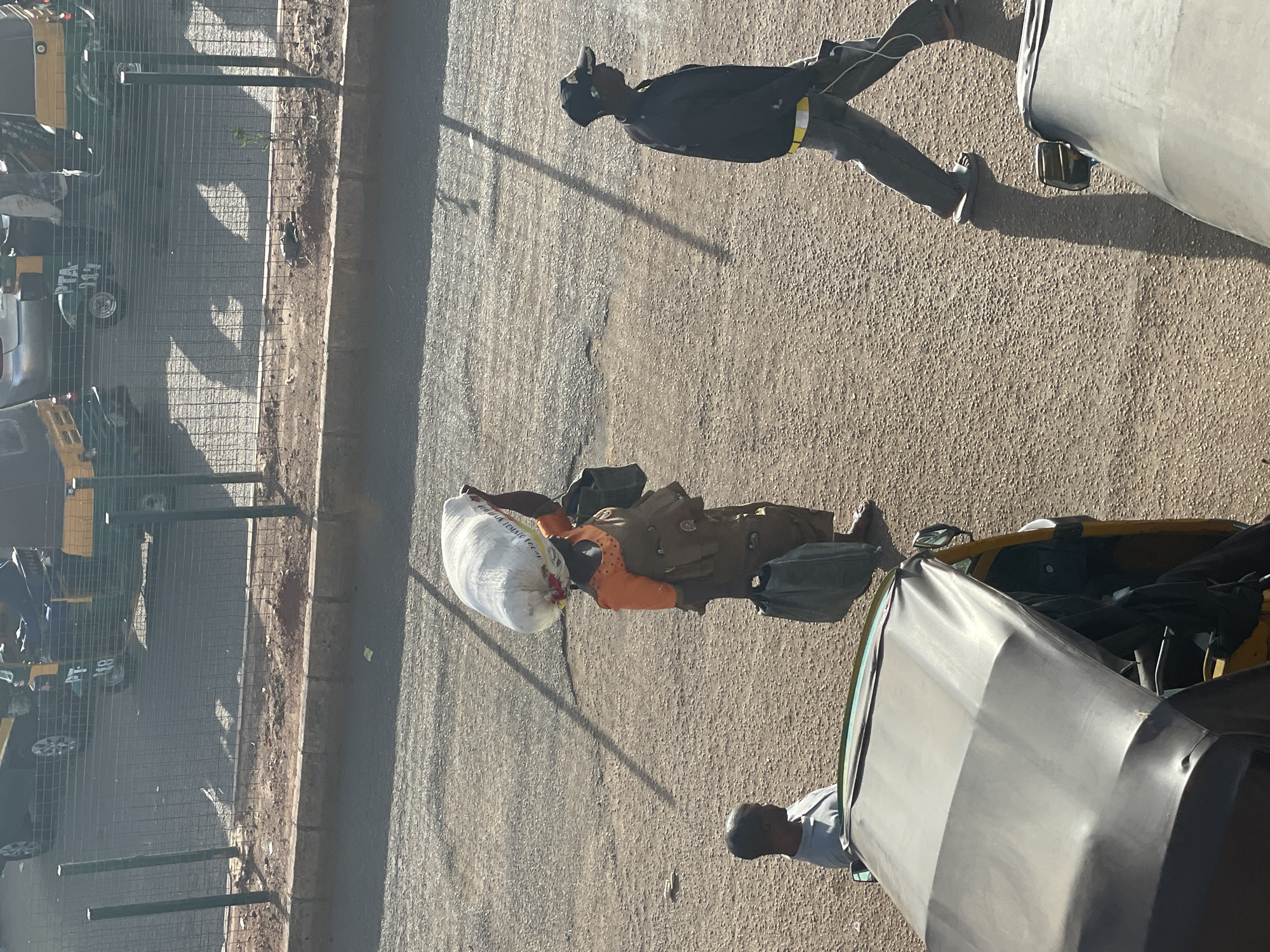 The image depicts a street scene in Bukuru, Nigeria. A woman carrying a large white sack walks along the side of a road adjacent to a wire fence, behind which are parked vehicles resembling tuk-tuks. The woman is wearing an orange shirt and what appears to be a brown outfit. Further down the road, a person in a black outfit with reflective striping is walking. A third person is standing closer to the camera behind what appears to be an auto rickshaw. The road surface is rough and textured. The light suggests it is daytime.