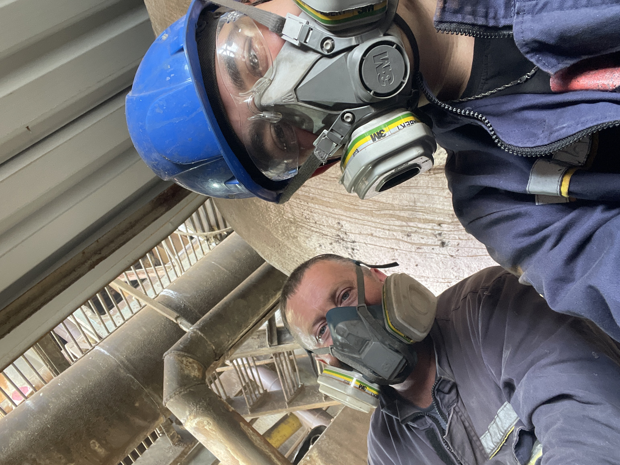 Two men are wearing industrial protective gear including respirators and safety glasses, with one also wearing a blue hard hat. They are in an industrial setting with large pipes and metal structures visible in the background. The lighting suggests an indoor environment. The image is a selfie, taken from a low angle.