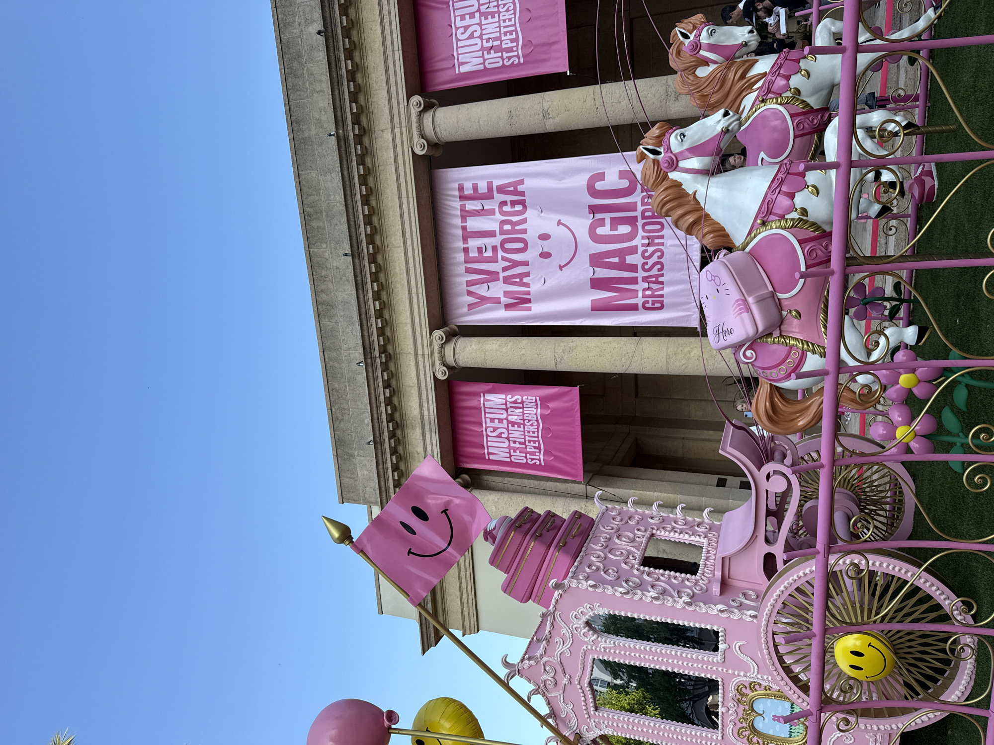 The scene depicts an outdoor art installation positioned in front of the Museum of Fine Arts in Saint Petersburg, Florida, United States. A large, ornate, pink and gold Cinderella-style carriage dominates the foreground. The carriage features intricate scrollwork, a yellow smiley face on its visible wheel, and a stack of pink suitcases on its roof. A pink flag with a black smiley face is affixed to a pole extending from the carriage, alongside large pink and yellow balloons.
Two white, life-sized carousel-style horses, adorned with pink harnesses, saddles, and gold accents, are positioned as if pulling the carriage. One horse carries a pink saddlebag featuring a "Hello Kitty" design. The entire display rests on artificial green grass and is enclosed by a decorative pink metal fence with gold scrollwork and pink flower motifs with yellow centers.
In the background stands a classical architectural building identified as the Museum of Fine Arts. It features a light-colored stone facade and prominent fluted Doric columns supporting an entablature. Three large pink banners hang vertically from the building. The leftmost and rightmost banners display "MUSEUM OF FINE ARTS ST. PETERSBURG." The central banner reads "YVETTE MAYORGA MAGIC GRASSHOPPER," incorporating a stylized smiley face and closed eyes. Several indistinct individuals are visible in the background, positioned behind the art installation near the museum entrance. The sky above is clear and blue, indicating bright daylight conditions.