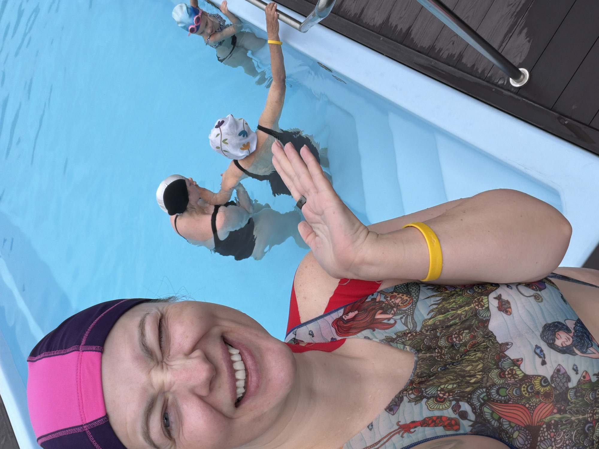 The image shows a group of women in a swimming pool. One woman, wearing a pink and purple swim cap and a swimsuit with a mermaid design, is in the foreground, smiling and raising her hand towards the camera. Behind her in the pool, there are two other women wearing swim caps and black swimsuits. One is reaching towards the pool's edge, where a metal handrail is visible on a dark wooden deck. Another woman is further away in the pool. The water is light blue, and the surrounding environment appears to be an outdoor pool area, potentially located in Ждановіцкі сельскі Савет, Belarus.