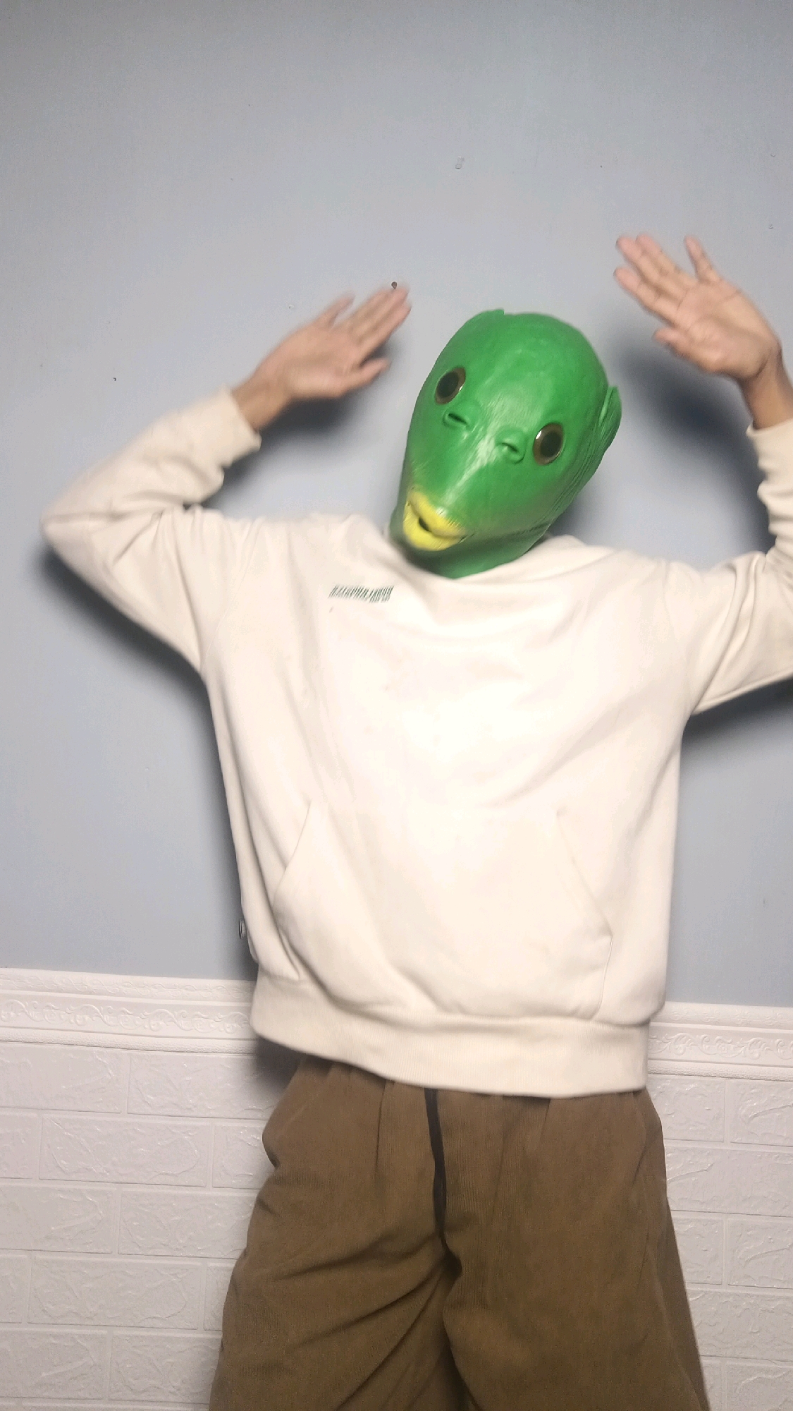 A person wearing a green alien mask and a cream-colored hoodie is standing in front of a light blue wall with a white textured baseboard. The person's hands are raised and slightly blurred, suggesting movement. They are also wearing brown corduroy pants. The background wall is plain, with some faint marks visible. The lower part of the wall has a distinct white brick pattern. The scene appears to be an indoor setting. There is no indication of the location being Kebumen, Indonesia.
