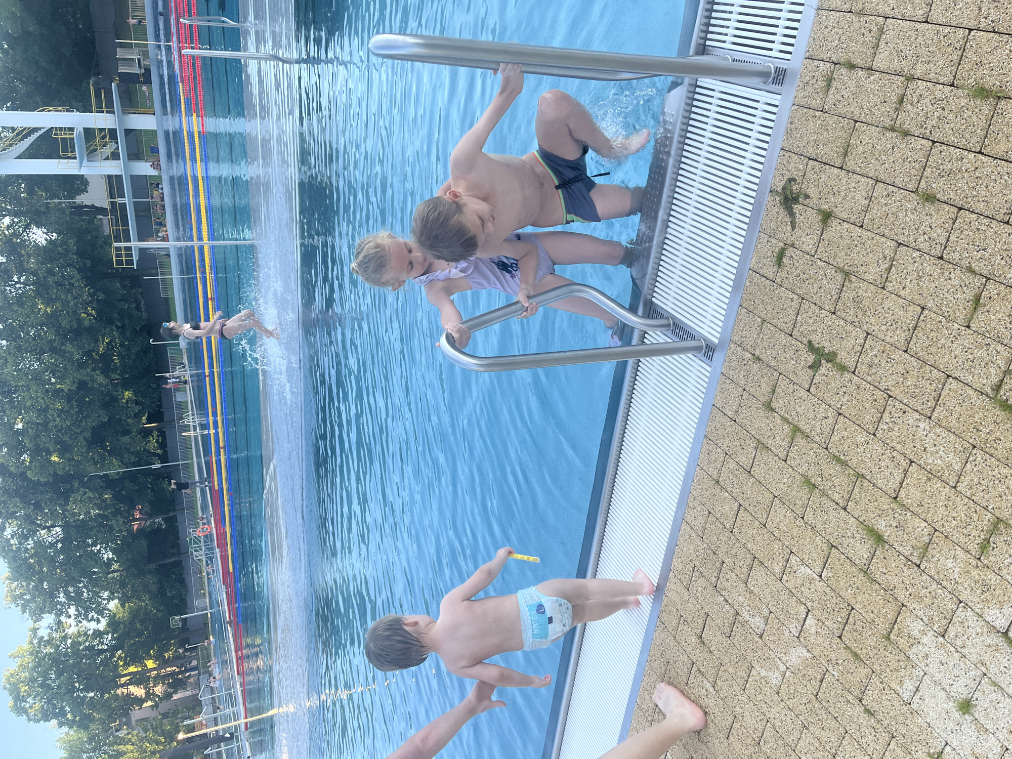 In a sunlit outdoor swimming pool area, a young child wearing a diaper stands at the edge of the pool, facing the water. To the right, a boy and a girl are using the pool steps. The boy is partially submerged in the water, holding onto a metal handrail, while the girl is also holding the handrail and looking down. In the background, other swimmers and a diving board are visible. The area around the pool is paved with light-colored interlocking bricks, and surrounded by green trees.