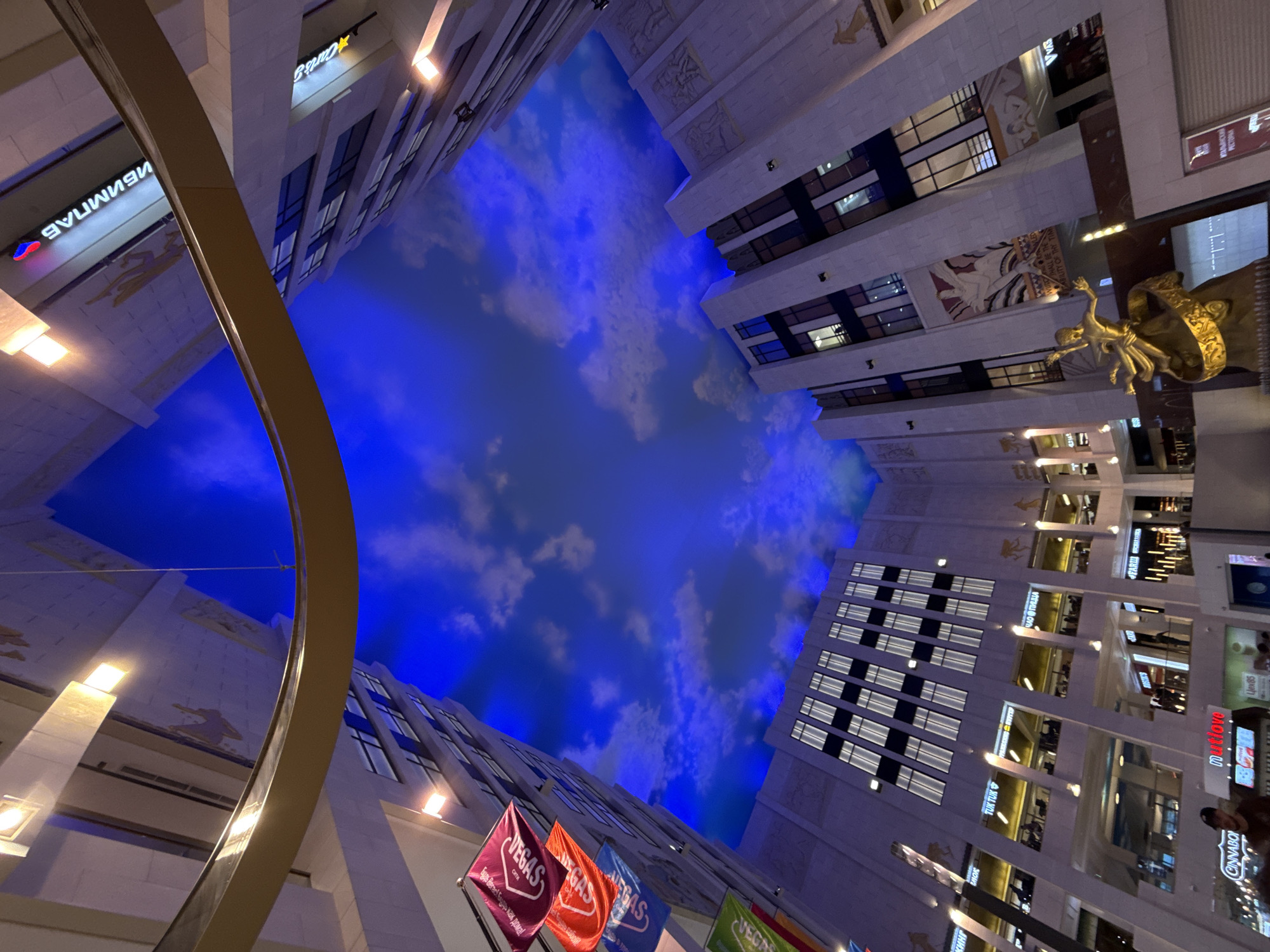 This image shows an interior view of a shopping mall, looking upwards towards a brightly lit ceiling that mimics a blue sky with clouds. The architecture of the mall is modern, with multiple levels of shops and walkways visible. Some shops have illuminated signs displaying brand names like "Carts &", "TUK TUK", "muutiove", "FARIMA", and "CINNABOR". There are also flags visible with the word "VEGAS" on them, suggesting a themed area or an event.
The perspective is wide and angled, making the buildings appear to stretch upwards. The lighting is predominantly artificial, with warm lights illuminating the shop fronts and bright, cool lights creating the sky effect on the ceiling. The overall impression is one of a vibrant and somewhat futuristic commercial space.
The image contains the following visible text: "VEGAS", "Сити", "MEGAS", "SHOWE", "CINNABOR", "TUK TUK", "ЧАО ПИЦ", "ПИЦЦА", "muutiove", "Цех 85", "FARIMA", "Carts &", "ИБИМПАБ", "LITY OF THY TIM", "ИТАЛЫНСКОЙ", "PECTOPAN", and "VVAS".
The activity appears to be a regular day in a shopping mall, with people visible in some of the shop windows, though they are small and indistinct. The setting is indoors, designed to evoke an outdoor sky, suggesting a controlled environment regardless of the time of day or external weather conditions. There are no strong emotional cues conveyed, beyond a sense of visual stimulation and commercial activity.