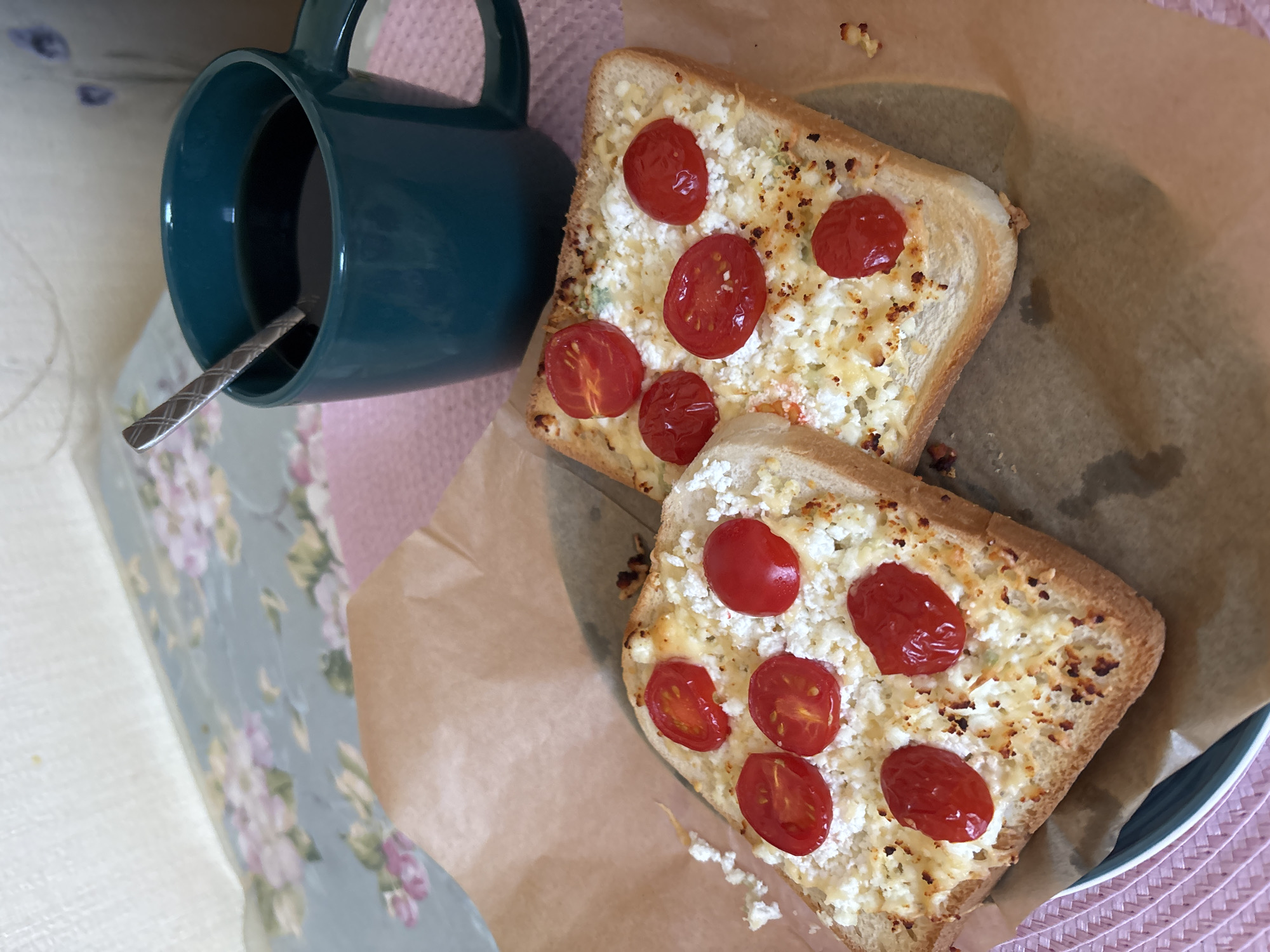 The image shows two pieces of toast topped with crumbled cheese and sliced cherry tomatoes. The toast is golden brown and appears to be toasted on both sides. The cheese is white and crumbly, and the tomatoes are bright red and appear to be cooked or warmed. A dark liquid, likely coffee, is in a teal mug with a silver spoon in it. The mug is placed on a pink placemat. The background is blurred but appears to be a table with a floral tablecloth. The image is shot from a slightly overhead angle. There is no indication of the city of Naberezhnye Chelny, Russia, in the image.