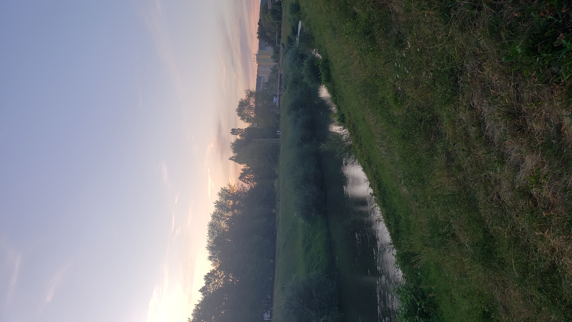 The image depicts a serene riverbank scene at dusk in Prešov, Slovakia. The sky transitions from a pale lavender near the horizon to a soft blue overhead, with faint wisps of clouds catching the last rays of sunlight. A dense line of trees, primarily deciduous, borders the far bank of a calm, dark river. Their silhouettes are etched against the twilight sky, with hints of orange and pink light filtering through the leaves.
In the middle ground, lush green grass slopes gently down to the water's edge. Scattered bushes and smaller trees punctuate the grassy expanse. Further in the distance, on the right side of the frame, several buildings, including apartment blocks, are visible, suggesting the presence of urban development near the natural landscape. A bridge crosses the river in the mid-distance, and a few small figures, likely pedestrians, can be observed near the river or on the far bank. The foreground is dominated by a steep, grassy embankment, overgrown with a mix of green and dry grasses, suggesting a wilder, less manicured area. The overall impression is one of tranquility and the gentle encroachment of nature into an urban environment.
