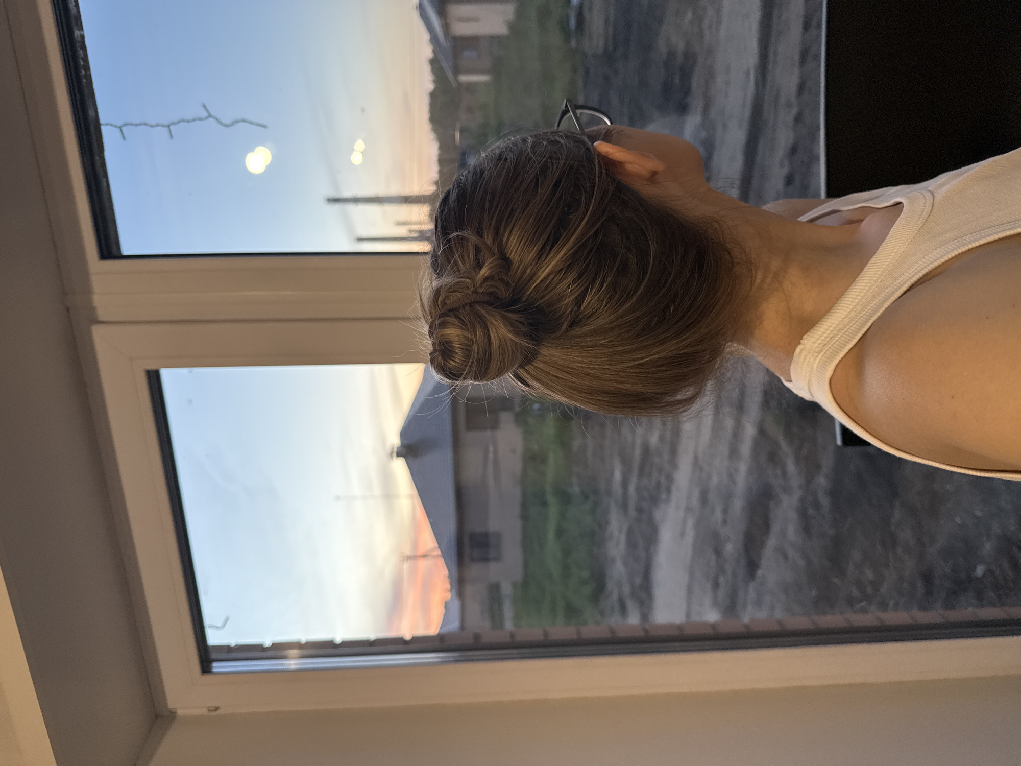 A woman with brown hair in a bun is looking out of a window. She is wearing glasses and a white tank top. The window is a white framed window. Outside the window is a view of buildings, trees, and sky. The sky is a combination of blue and orange, suggesting it is either sunrise or sunset. The location is potentially in Chaikovsky, Russia, based on the prompt. The image is taken from an interior perspective.