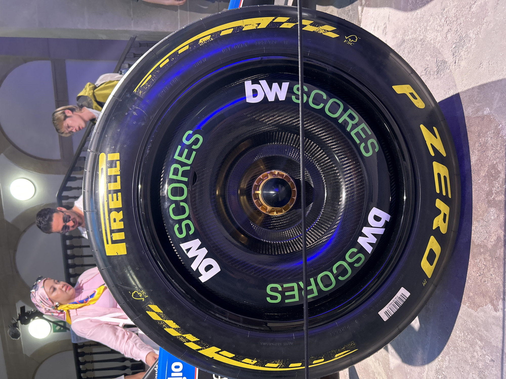 The image shows a close-up of a Formula 1 tire, with part of a carbon fiber wheel visible. The tire has yellow lettering that reads "P ZERO" and "PIRELLI". Green lettering on the tire reads "bwSCORES" repeated twice. There are also yellow checkered patterns on the side of the tire.
In the background, three individuals are visible. One is a woman wearing a pink headscarf and a pink jacket over a yellow shirt. To her right, a man wearing sunglasses and a white t-shirt is partially visible. Further back and to the right, a young person with light-colored hair is wearing a yellow jacket.
The setting appears to be indoors, with arched architecture and lighting that suggests a public space. The flooring is a light-colored textured surface. Based on the visible architecture and lighting, this scene could be located at the Fira de Barcelona Gran Via exhibition center, a common venue for major events and product launches in Barcelona, Spain.