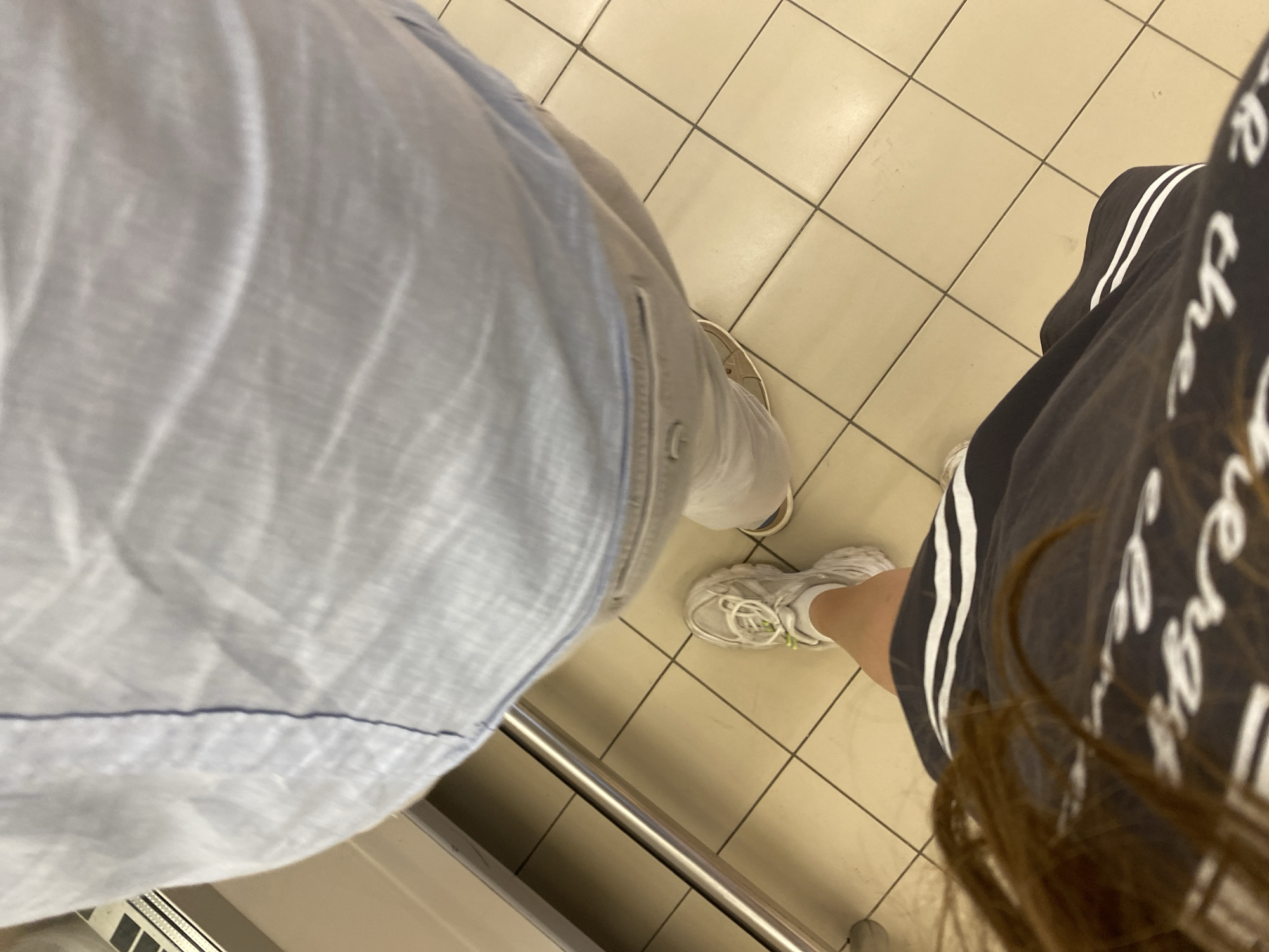 The image shows a low-angle view of two people's lower bodies and legs, standing on a tiled floor. One person, wearing a light blue button-up shirt and grey shorts, is visible from the waist down. Their shorts have a pocket with a button. Another person is in the foreground, wearing a dark skirt with white stripes and white sneakers. They have long brown hair. The background features a metal railing and a white surface, possibly a counter or shelf. The floor is made of square, light beige tiles with dark grout lines. The scene appears to be indoors, potentially in a commercial or public setting. The location is stated to be in Kuzmolovskiy, Russia.