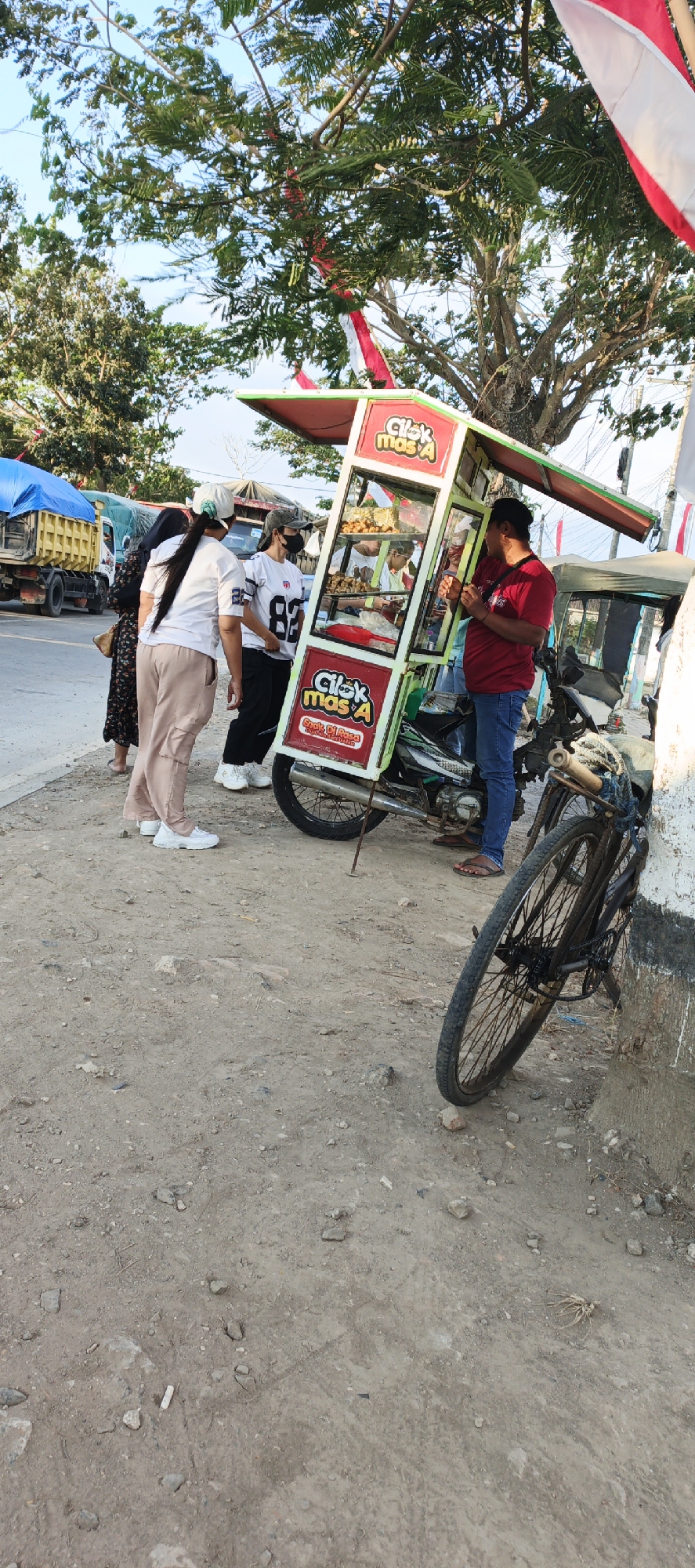 In Kasiyan Timur, Indonesia, a street food vendor's cart, adorned with "Cilok Mas A" branding and red and white flag accents, is positioned on a dirt roadside. A man in a red t-shirt and blue jeans attends to the cart, interacting with customers. Two women in casual attire stand nearby, observing. One woman wears a white t-shirt and beige pants, while the other has a white t-shirt with a black "82" graphic and a dark baseball cap. A third woman, partially obscured, stands further left, wearing a patterned dress and dark headscarf. The cart, mounted on a motorcycle chassis, displays various food items within its glass enclosure. A black bicycle is parked leaning against a textured, striped wall to the right of the frame. In the background, a yellow dump truck and other vehicles are visible on a paved road, suggesting an active urban environment. Trees with abundant green foliage and clear blue sky complete the scene.