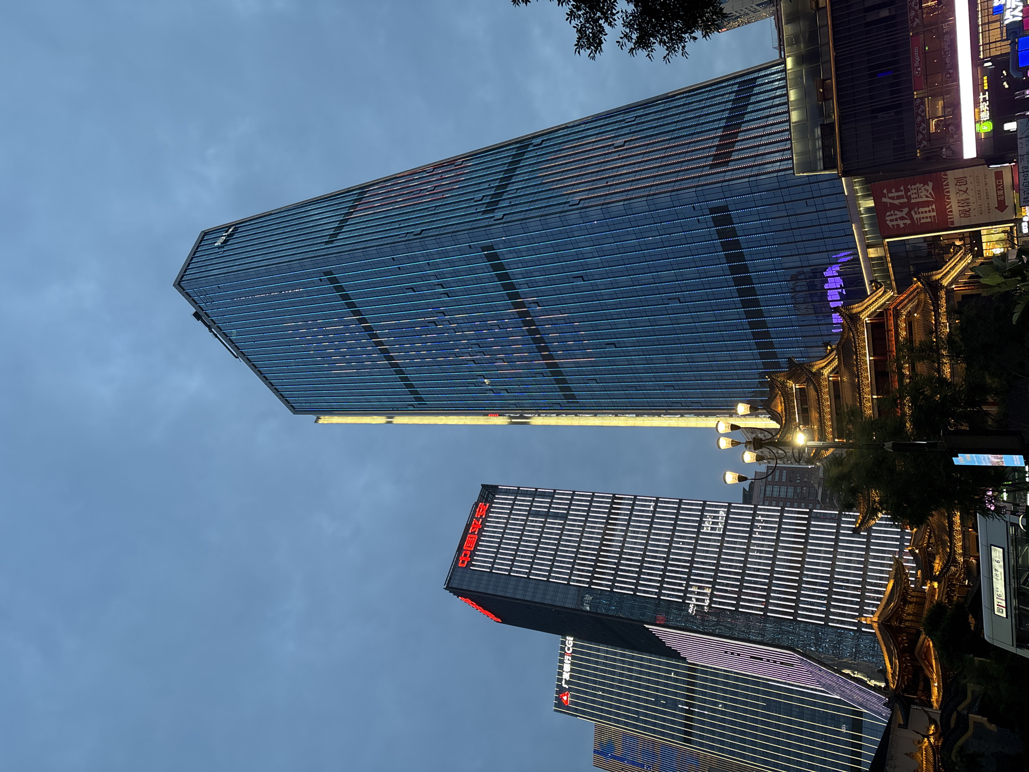 The image depicts a nighttime urban scene in Yuzhong District, Chongqing, China. In the foreground, a multi-tiered, traditional Chinese architectural structure with illuminated eaves is visible. In front of this structure, a white bus with a digital display indicating routes is parked. To the left of the bus, a street light with multiple lamps illuminates the scene.
Dominating the background are several modern skyscrapers. The tallest building on the right is a blue glass skyscraper adorned with vertical lines of colorful LED lights. To its left, a darker skyscraper features red Chinese characters at its top, with vertical white lights illuminating its facade. Further left, another skyscraper displays vertical purple and white lights. Signs with Chinese text are visible on some of the buildings and on awnings below the skyscrapers, one of which reads "我在重庆" (I am in Chongqing). The sky is overcast and dark, suggesting it is either dusk or night. The overall impression is of a bustling, modern cityscape integrating traditional elements.