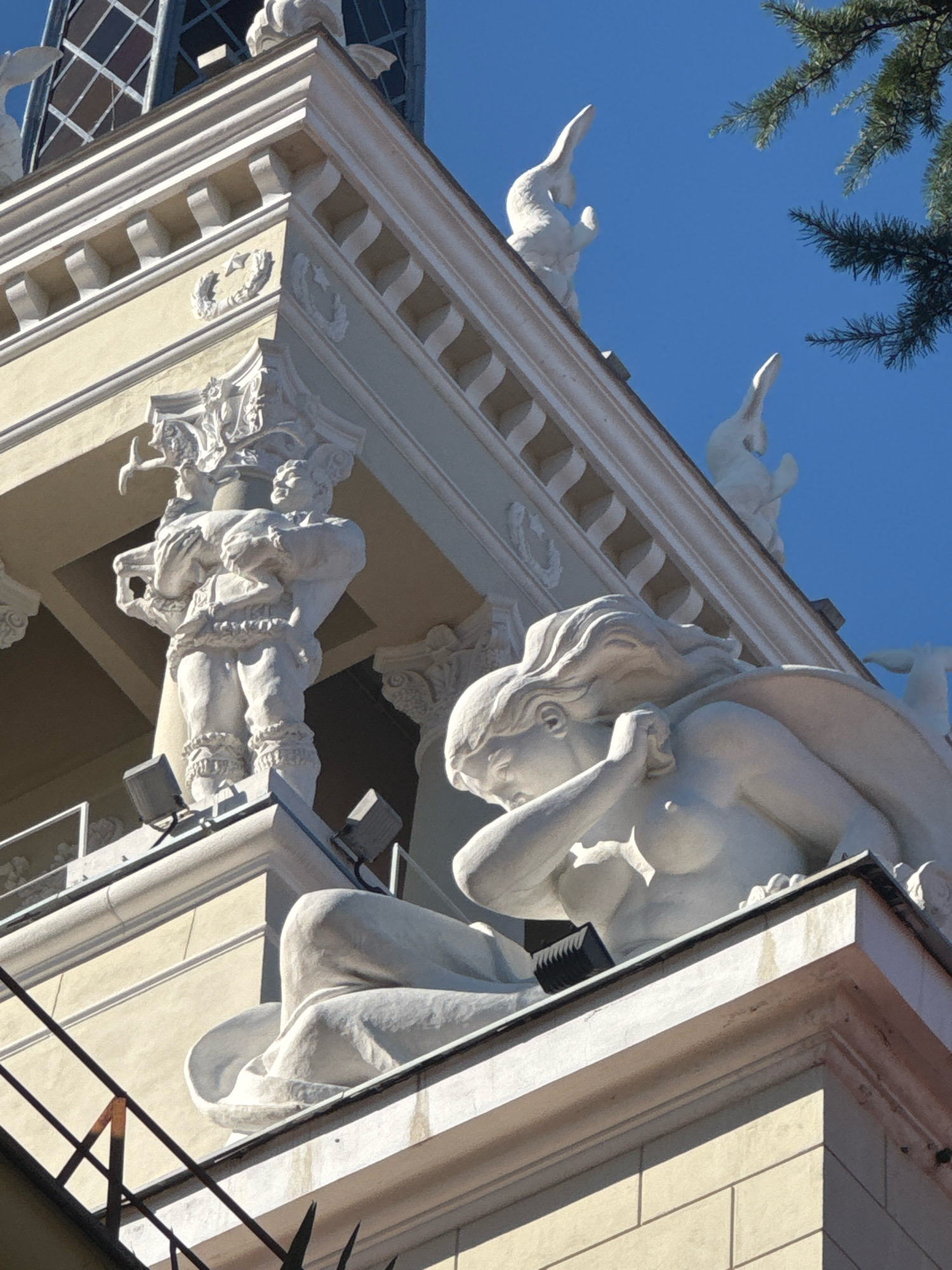 The image shows the exterior of a building in Sochi, Russia, with a clear blue sky as the background. The building's facade features classical architectural elements, including ornate cornices and decorative reliefs. Several white sculptures are visible on the building's upper levels. In the foreground, a large sculpture of a reclining nude woman is depicted, with her head turned and her arm raised to her face. Behind her, a sculpture of a muscular male figure holding a large object is partially visible. Further up, several stylized bird-like sculptures are perched on the roofline. A section of a pine tree branch is visible in the upper right corner of the image. The building appears to be adorned with spotlights and decorative metalwork. The overall impression is one of grand, artistic architecture set against a bright, sunny day.