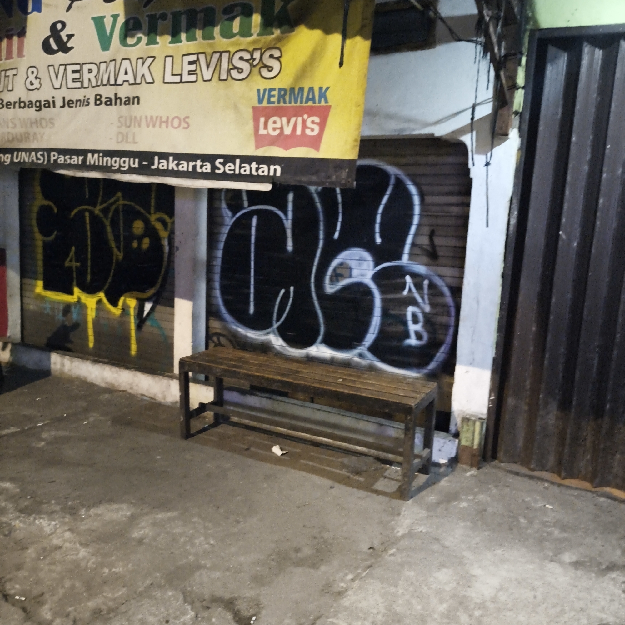 The image shows a street scene at night in South Jakarta, Indonesia. A weathered yellow banner hangs across the scene, displaying text in Indonesian, including "VERMAK LEVIS'S" and "VERMAK Levi's". The banner is partially obscured by graffiti on a rolling metal door behind it. To the right, a dark brown, ribbed metal door is visible. In the foreground, a wooden bench with a metal frame sits on a concrete ground that appears wet and dirty. The overall environment suggests an urban setting, possibly a shopfront or workshop, with a slightly dilapidated feel. No people are visible in the frame.