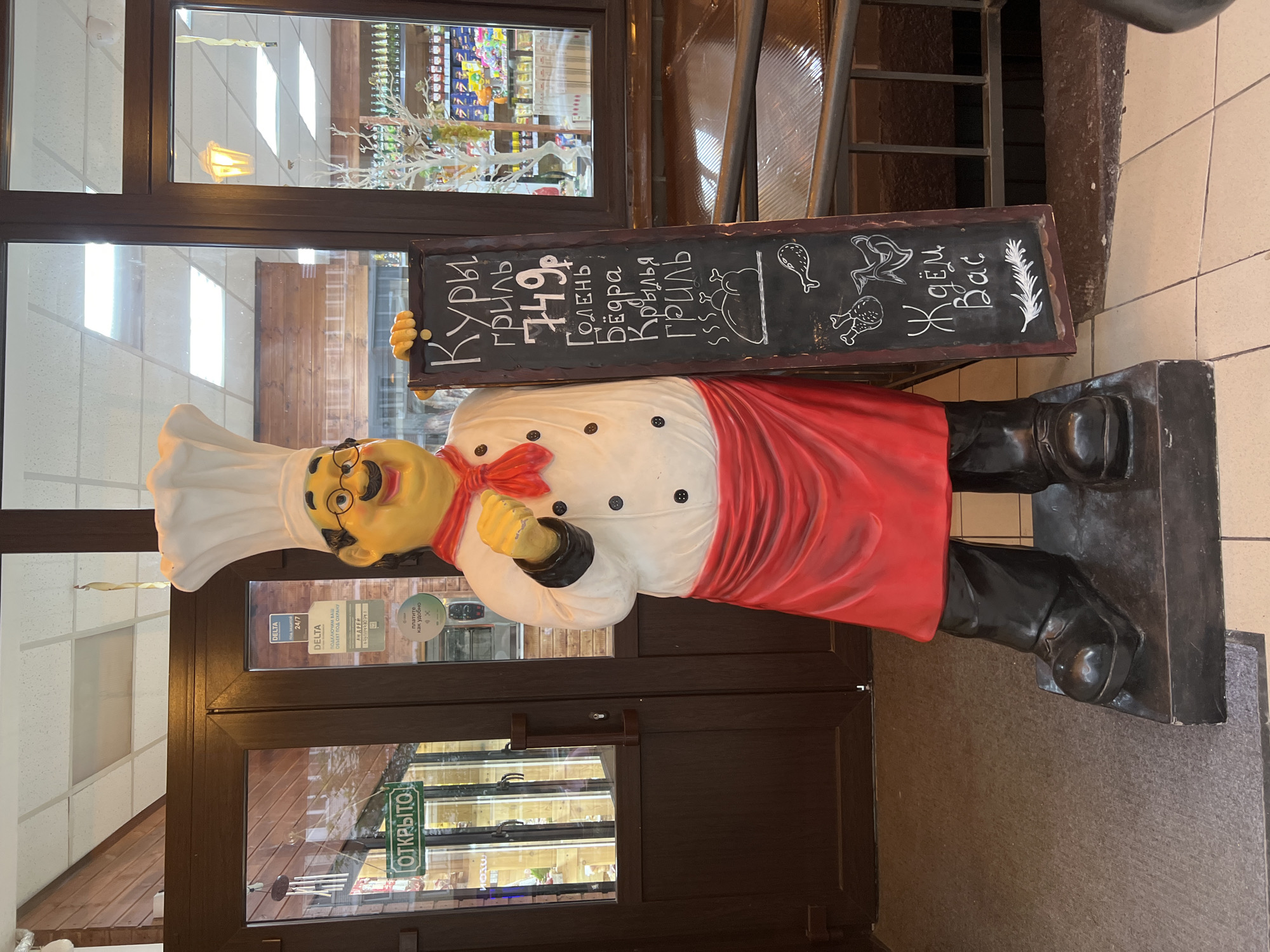 A life-sized, cartoonish chef statue stands facing forward, positioned directly in front of a double glass door entrance. The statue depicts a jovial figure with yellow skin, a black mustache, round spectacles, and is attired in a white chef's hat, a white double-breasted jacket with black buttons, a red ascot, a red apron, black trousers, and black boots. The chef holds a tall, narrow black chalkboard menu in its left hand, written in white chalk. The menu lists "Grilled Chicken" for "749 rubles," followed by options for "Drumstick," "Thighs," and "Grilled Wings," illustrated with chalk drawings of chicken. At the bottom, it reads "We are waiting for you."
Behind the statue, the double-leaf glass doors are framed in dark brown. The left door displays a green sign with the word "OPEN" in white Russian text. The right door is adorned with several informational stickers, including "DELTA 24/7" security service logos and contact information in Russian, and a "pay as convenient" payment method icon. Through the glass, a retail interior is visible, featuring shelves stocked with numerous bottles and packaged products, a decorative white tree, and a white suspended ceiling with fluorescent lighting. The floor beneath the statue consists of light-colored tiles, while the immediate foreground has a gray carpet. The statue rests on a dark, rectangular base. This scene is located in the city of Krasnodar, Russia.
