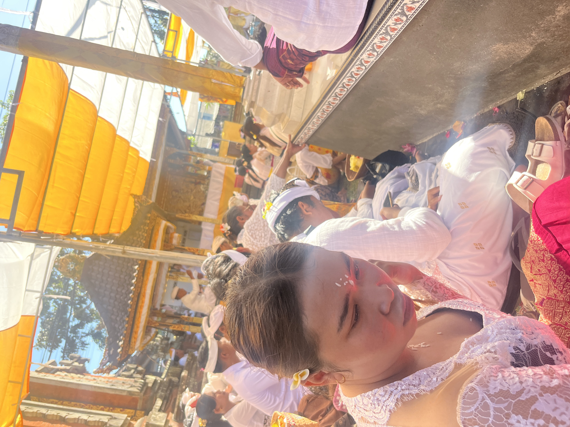 In Mendoyo, Indonesia, a group of people are gathered for a ceremony, likely a religious or cultural event, as indicated by their attire and the surrounding architecture. In the foreground, a woman with dark hair tied back is wearing a white lace top and a vibrant pink sarong. A small white flower is tucked behind her left ear, and a white mark adorns her forehead. She is looking down, with her chin resting on her hand, appearing contemplative. Rice grains are visible on her chest. Several other individuals, dressed in white attire with white headbands, are seated around her, some participating in the ceremony, others observing. Behind them, a temple-like structure with a dark, tiled roof and ornate details can be seen, topped with a green ornamental cap. The area is shaded by yellow and white awnings or tarps, suggesting an outdoor gathering. The ground is covered with dirt and scattered with small flower petals, further indicating the ceremonial nature of the event. In the background, more people are present, and glimpses of buildings and trees suggest a village or town setting. The overall atmosphere appears solemn and respectful.